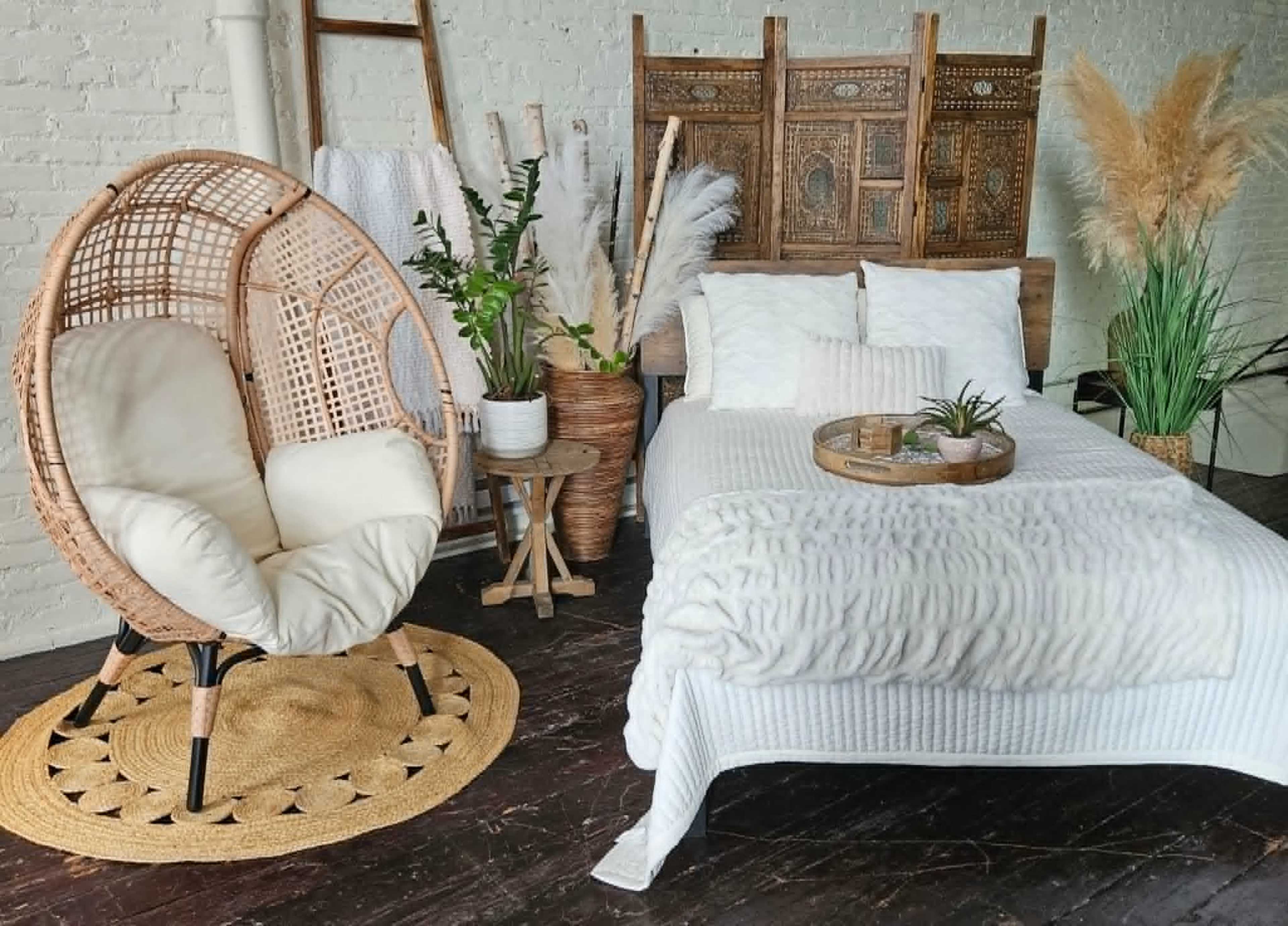 The image shows a cozy bedroom setup featuring a bed with a light-colored blanket, a rattan chair, and various decorative plants in woven baskets.