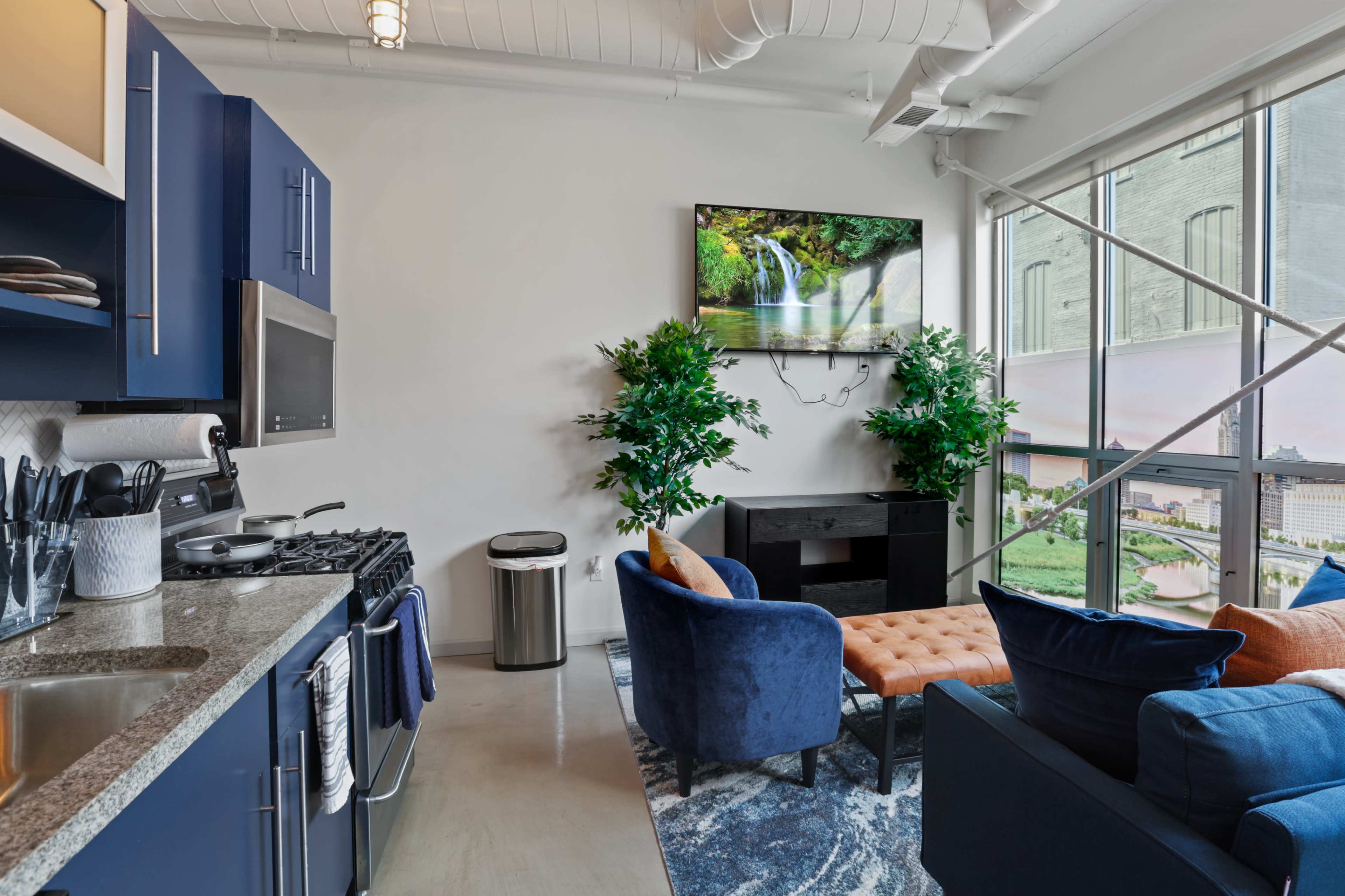A modern kitchen and living area feature dark blue cabinetry, a granite countertop, and a seating area with a tufted orange sofa beside a large window.