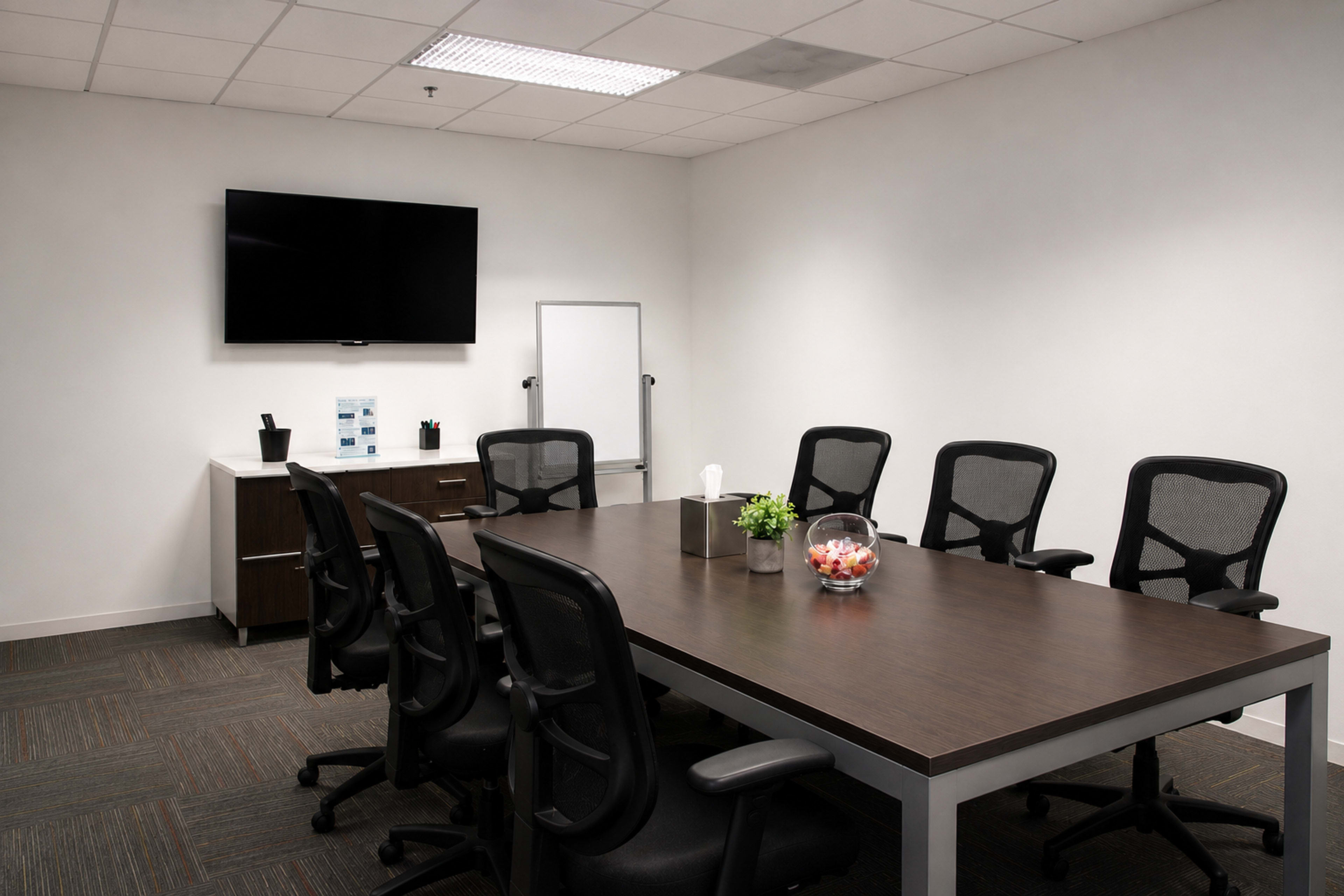 A conference room features a large wooden table surrounded by six black ergonomic chairs, a TV mounted on the wall, and a side cabinet with office supplies.