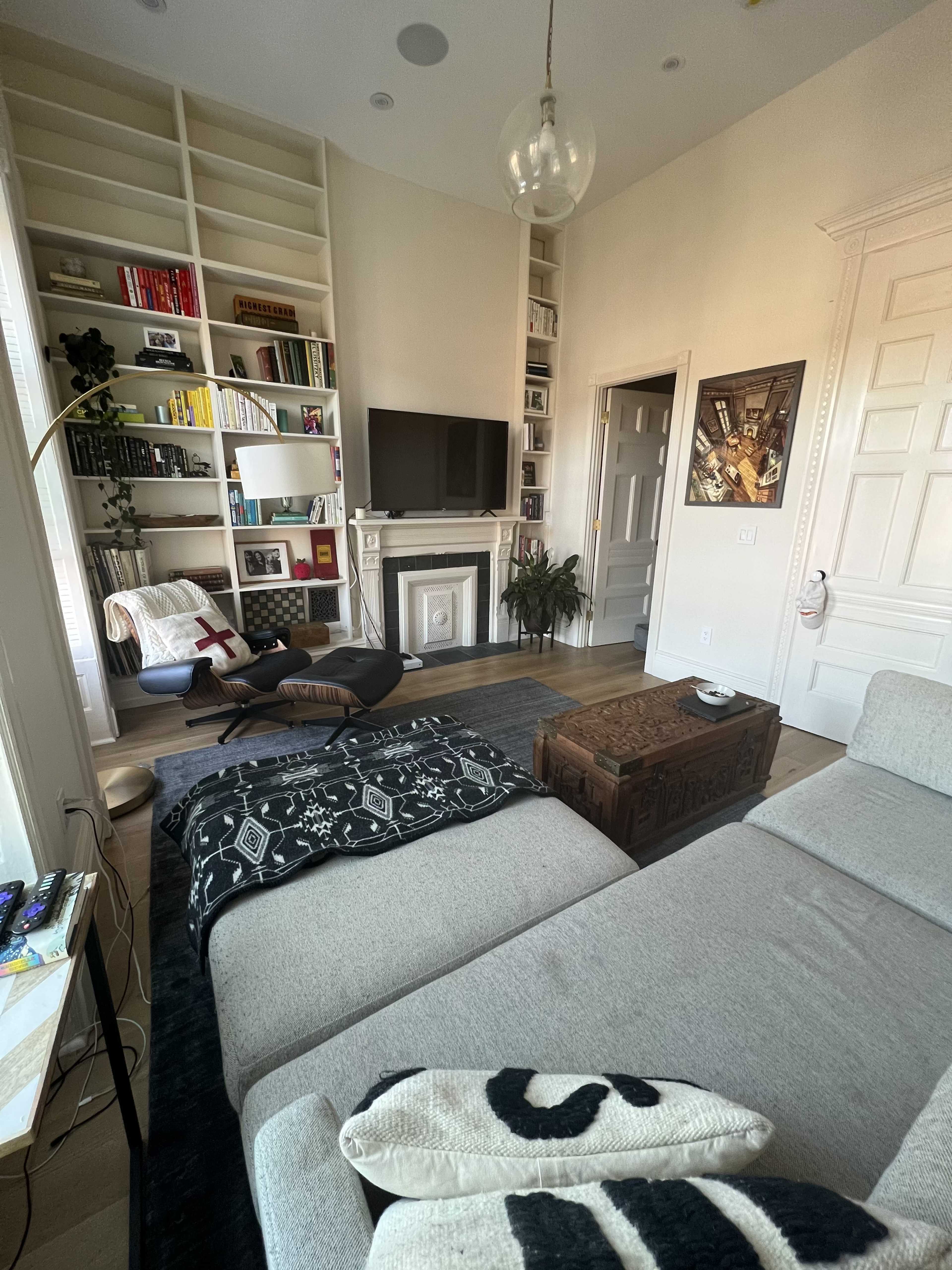 The room features a cozy living space with a sectional sofa, a coffee table, a bookshelf filled with books, and a television mounted above a fireplace.