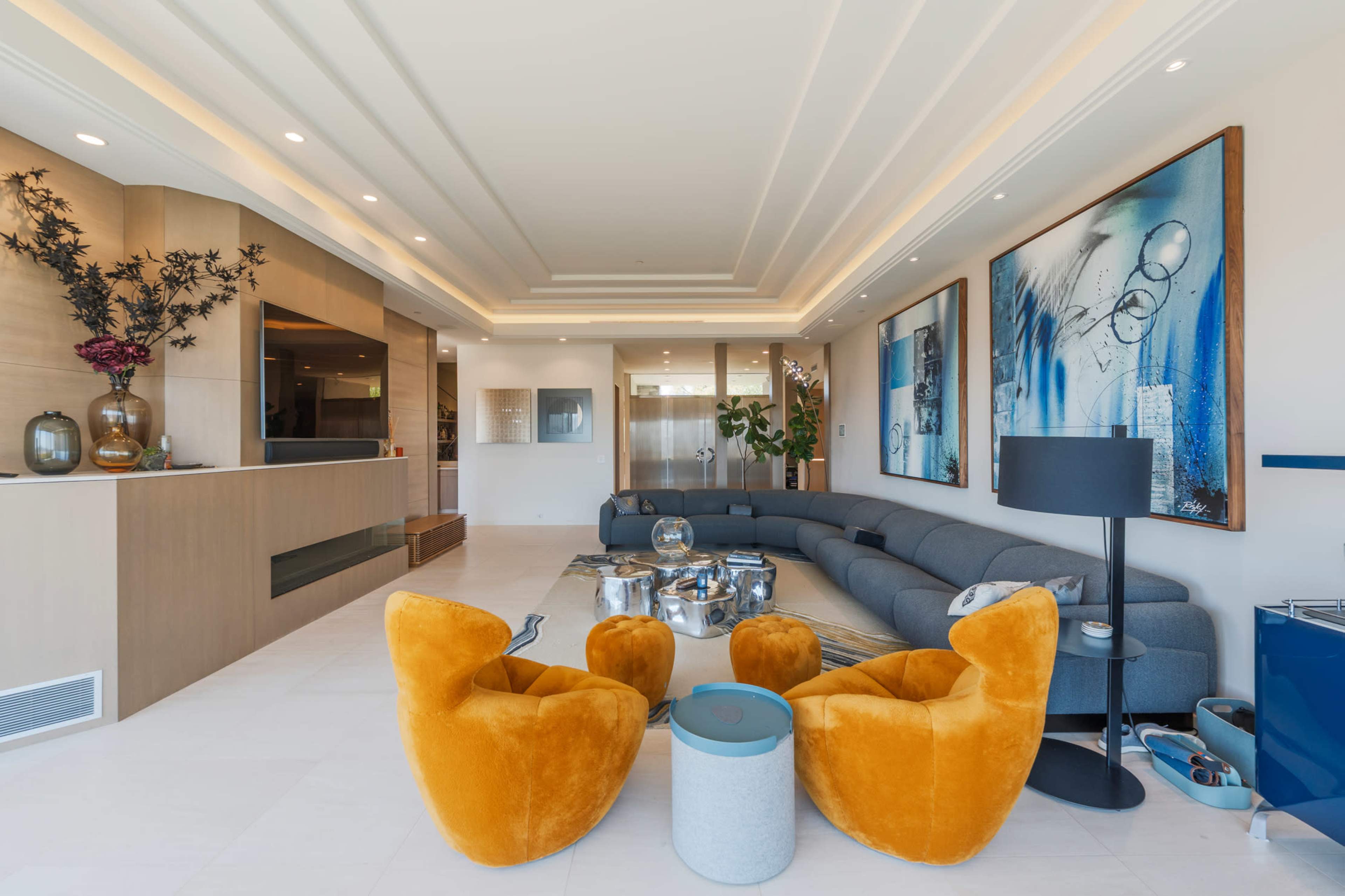 The image shows a modern living room with a curved gray sofa, two bright orange chairs, a coffee table, and large artwork on the walls.