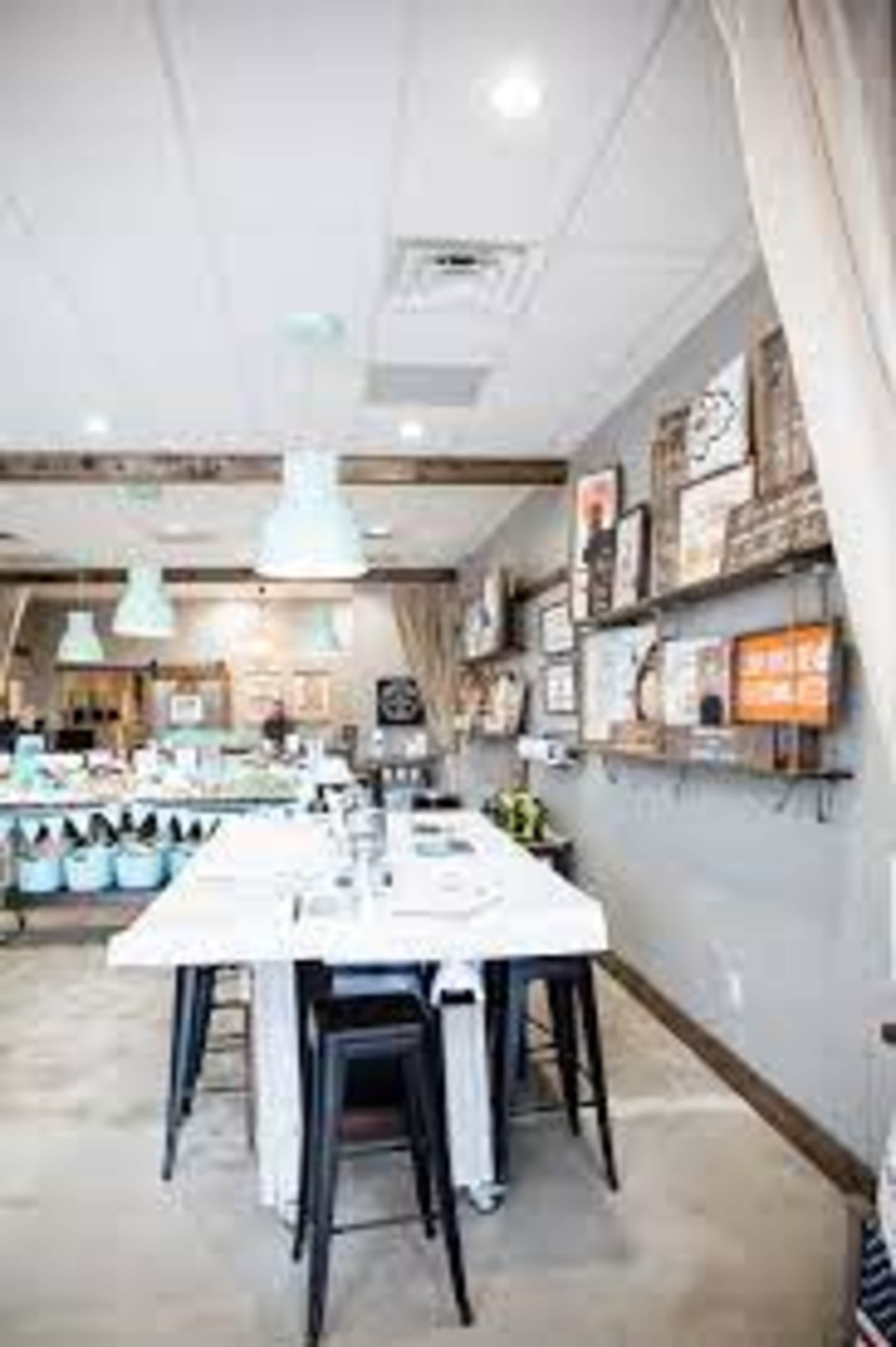 The image shows a modern café interior with a long white table, black stools, and walls adorned with various framed artwork.