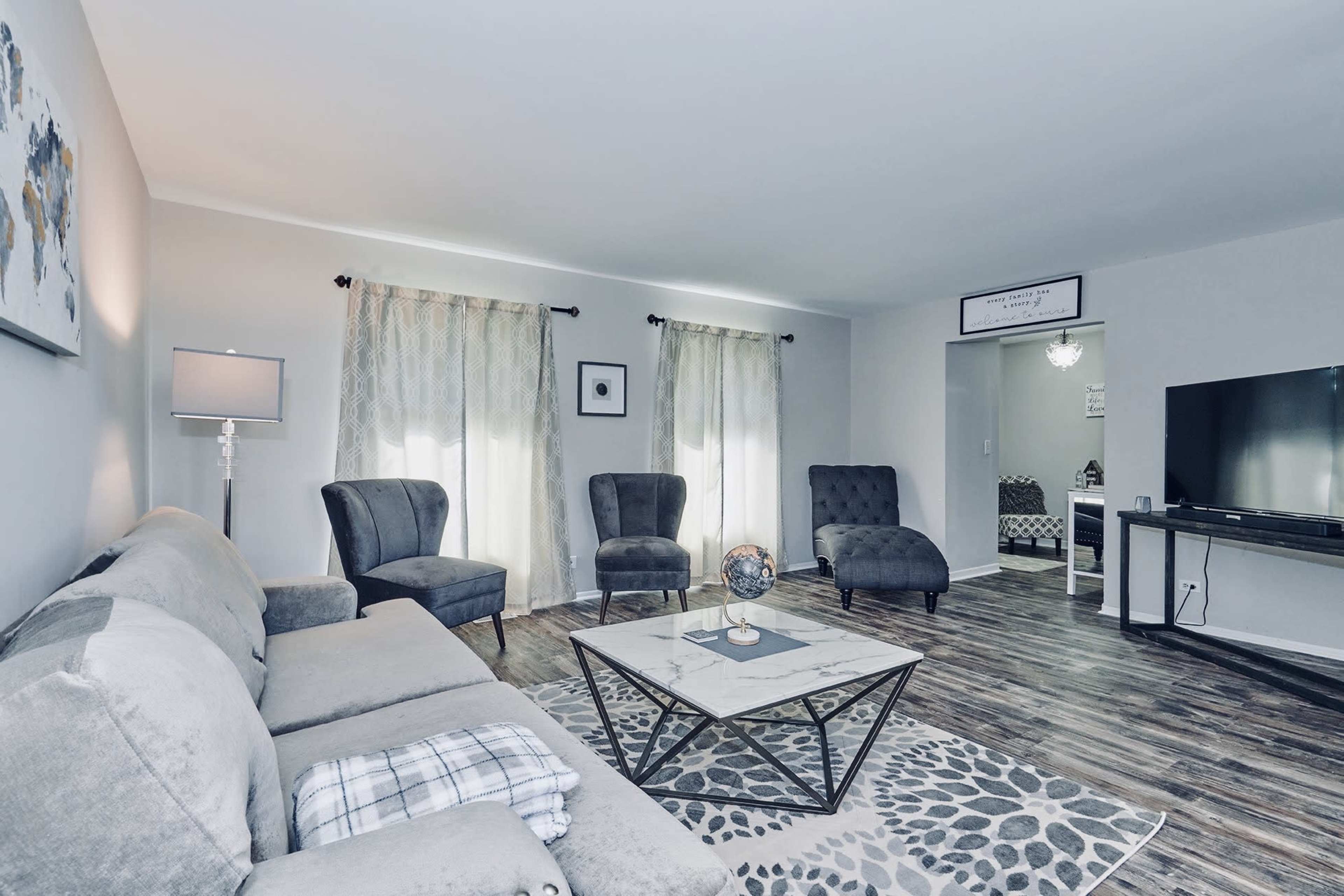 A living room with a gray sofa, two armchairs, a coffee table, and a television, complemented by light curtains and wooden flooring.