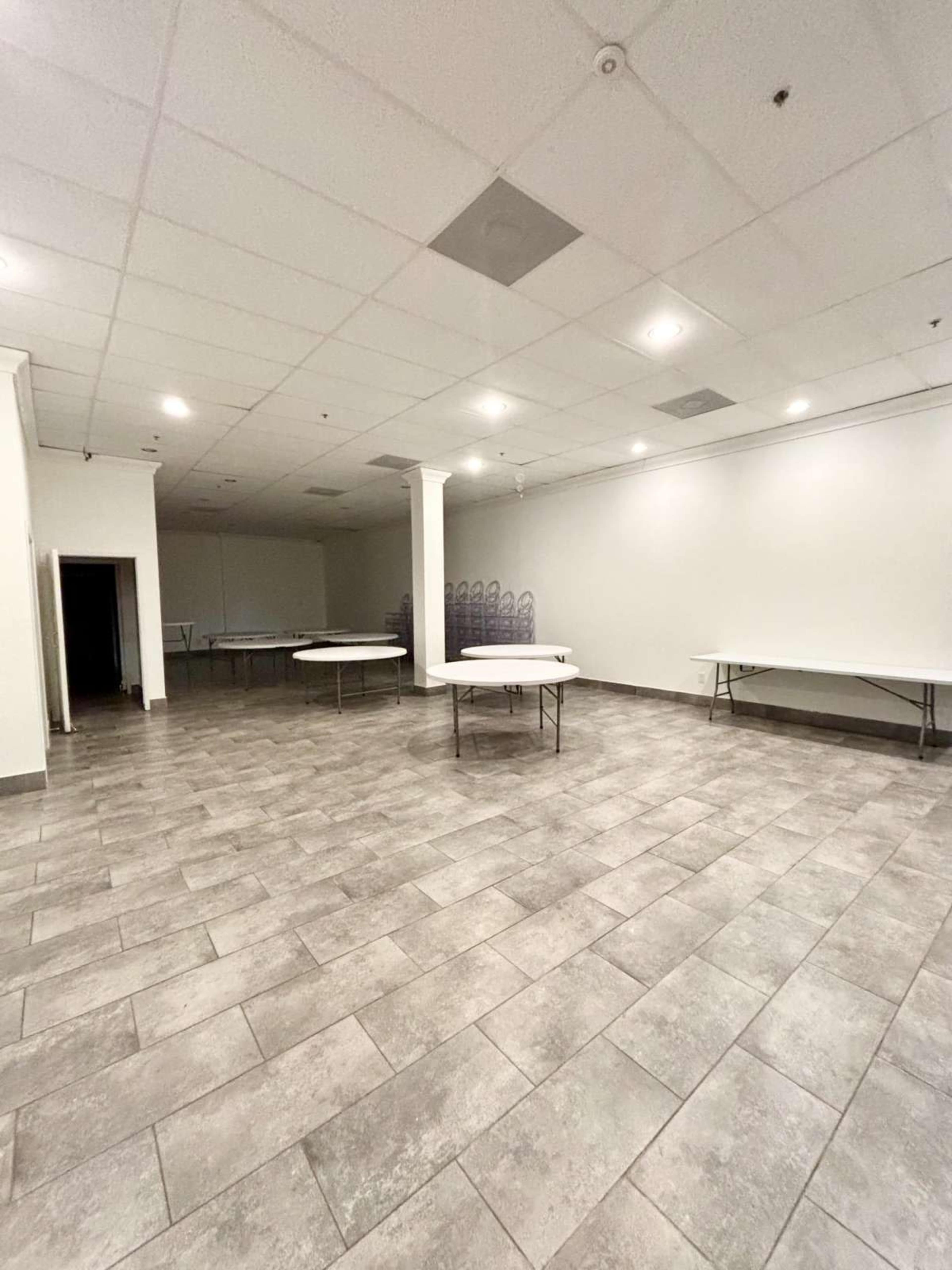 The image shows an empty room with gray tiled flooring, several folding tables arranged, and a stack of chairs in the background.