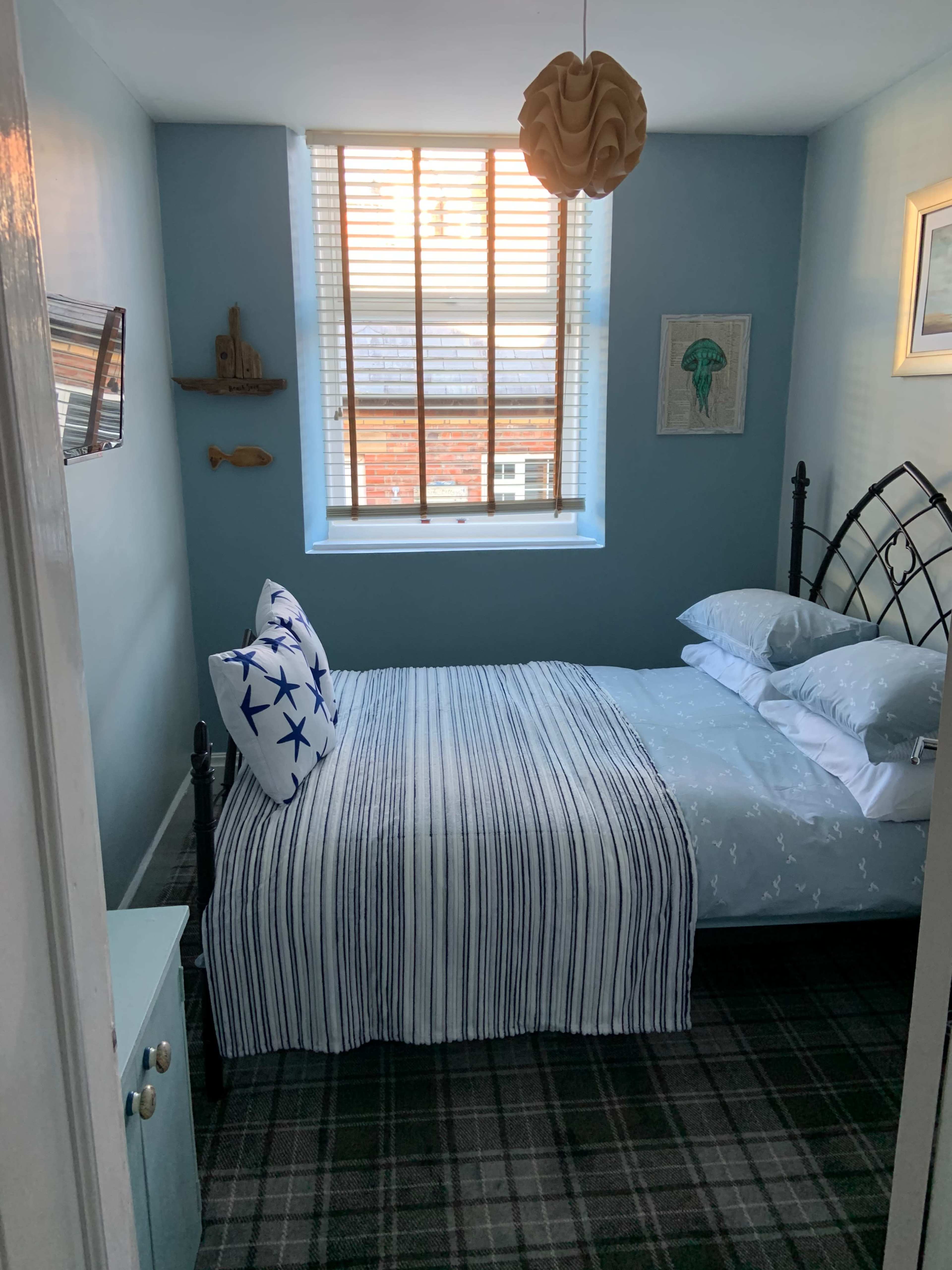 A small bedroom with a bed covered in a striped blanket, featuring decorative pillows, a window with blinds, and light blue walls.
