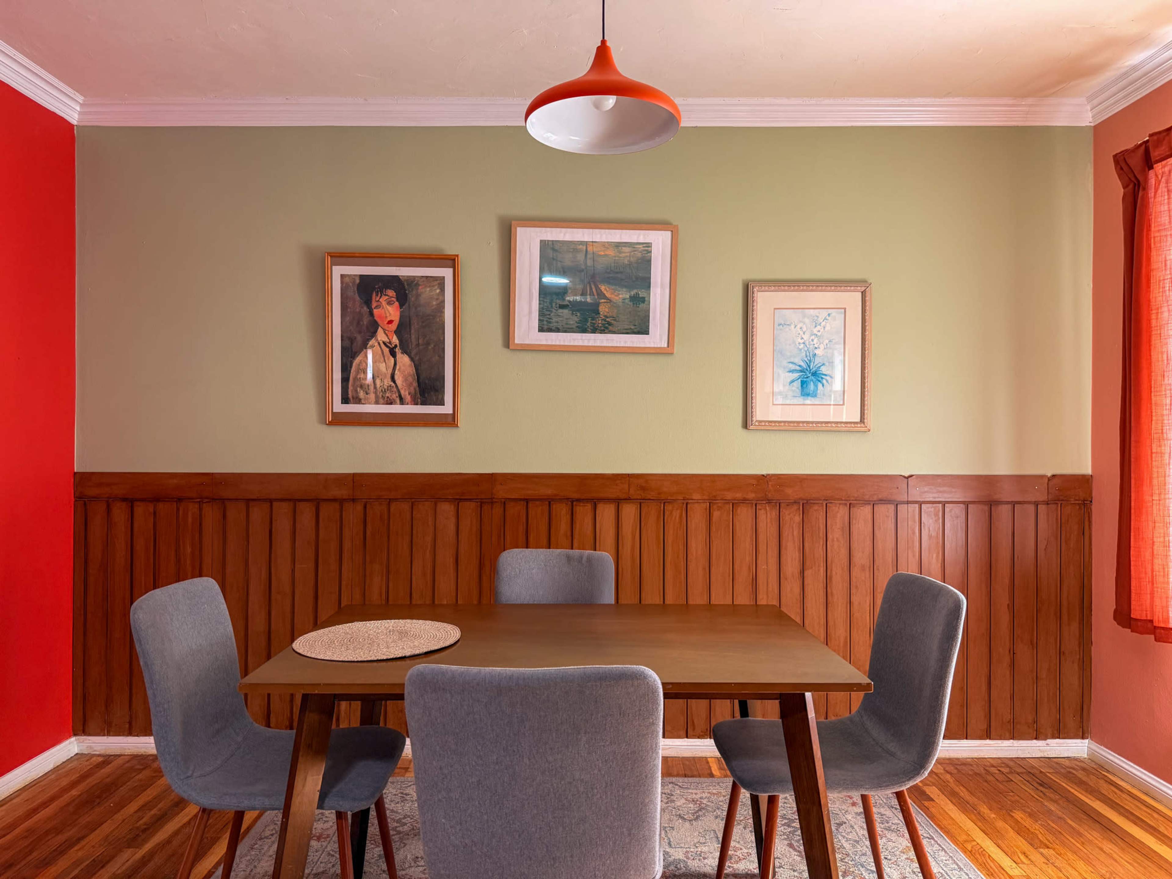 A dining area features a wooden table surrounded by four upholstered chairs, with framed artwork hanging on the walls and a pendant light above.