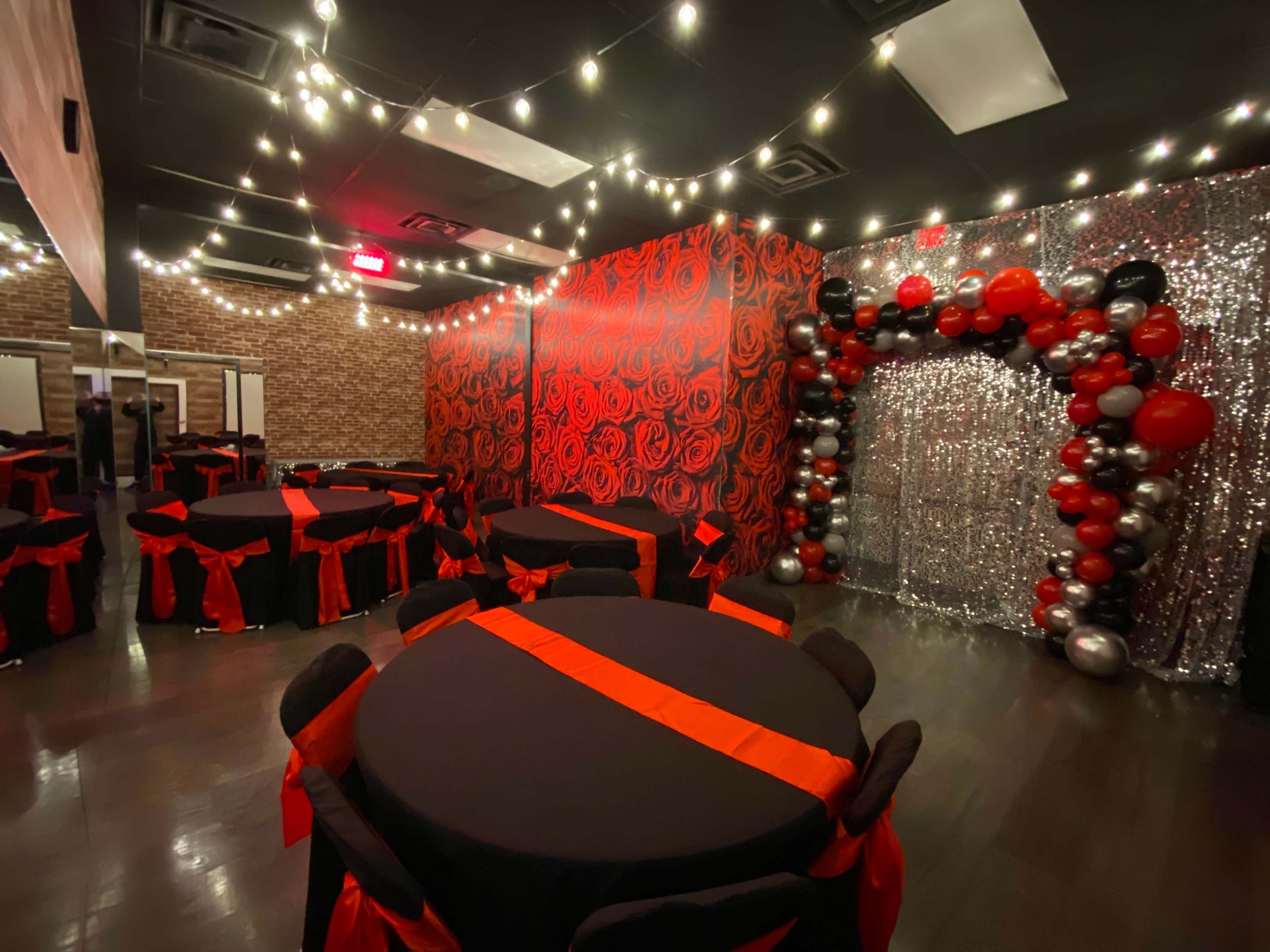 A decorated event space with round tables covered in black tablecloths and red accents, surrounded by a wall of red roses and a shimmering silver backdrop with a balloon arch.