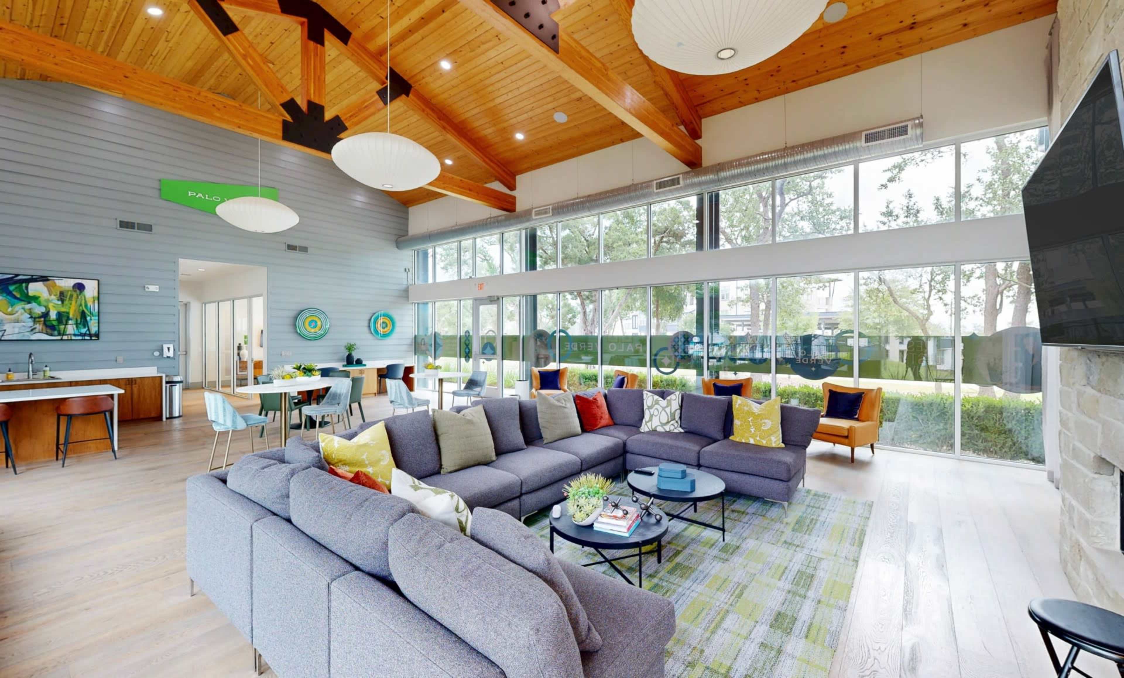 The image shows a modern communal living area featuring a large sectional sofa, a coffee table, and large windows that allow natural light to fill the space.
