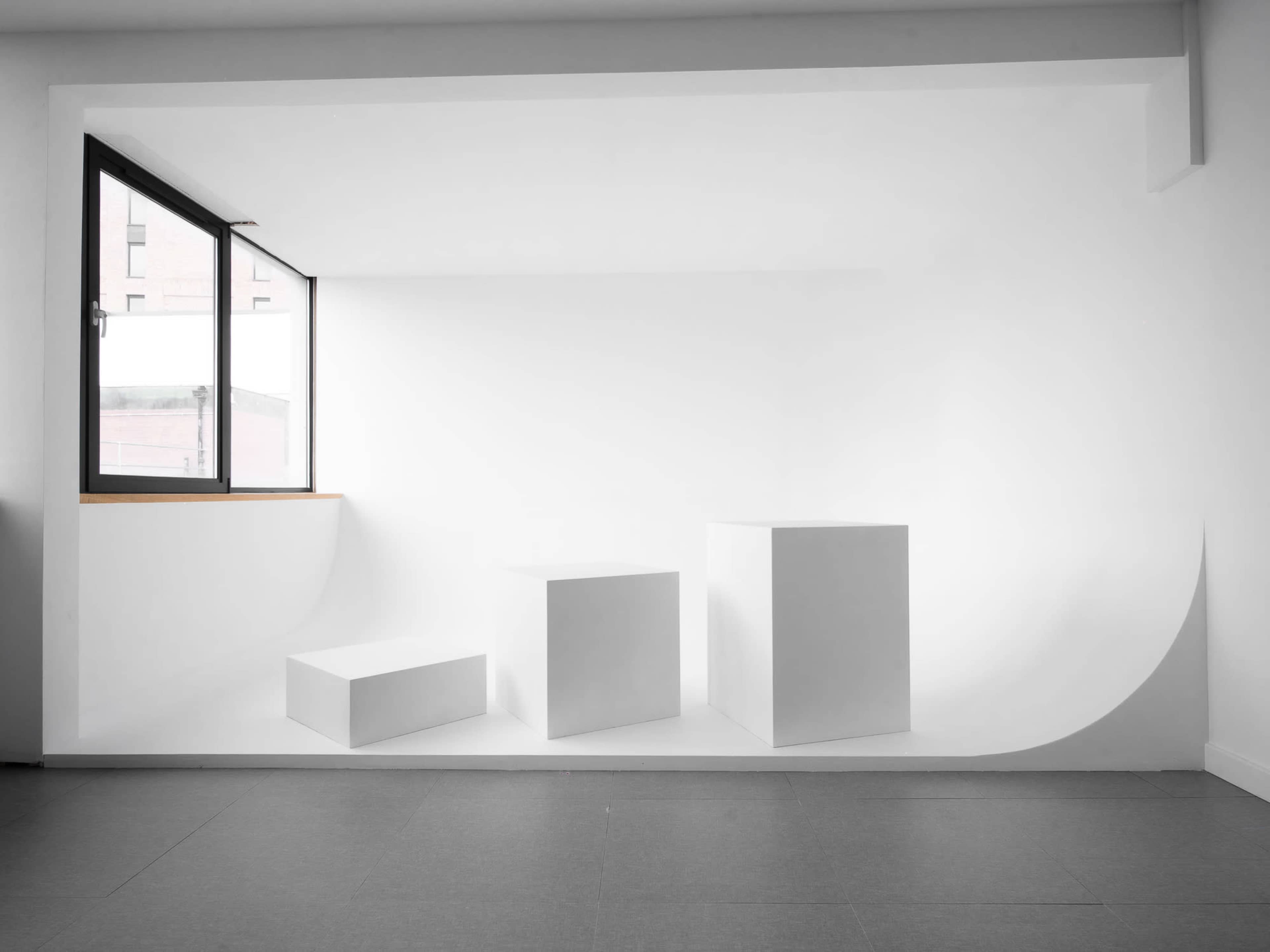 The image shows a minimalist interior space featuring three white geometric-shaped pedestals arranged against a white wall with a large window.