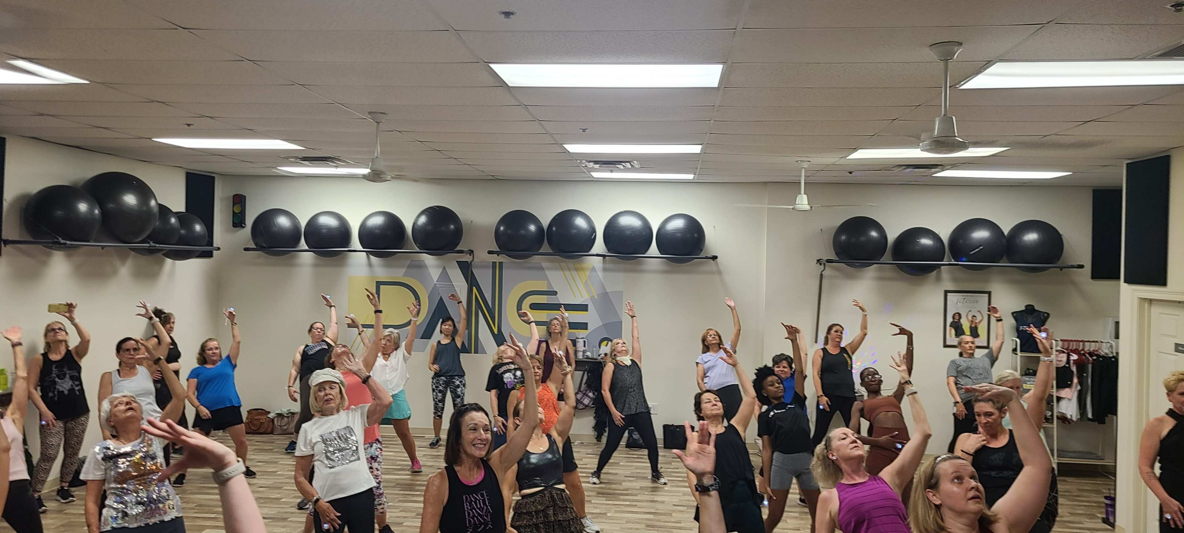A large group of people is participating in a dance class, raising their arms and following movements in a spacious studio with exercise balls on the walls.