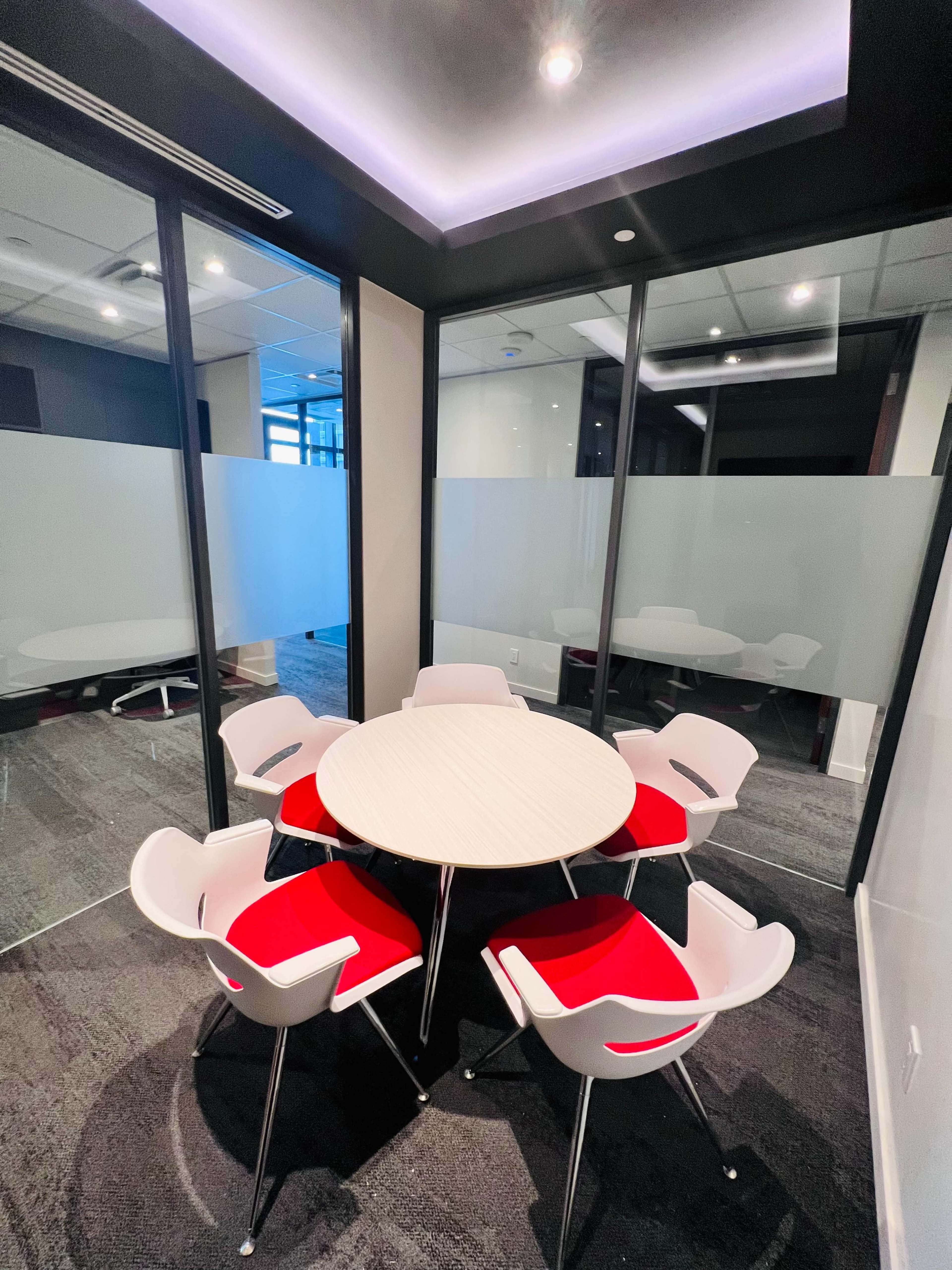 A small conference room features a round table surrounded by six chairs with red cushions, enclosed by glass walls.