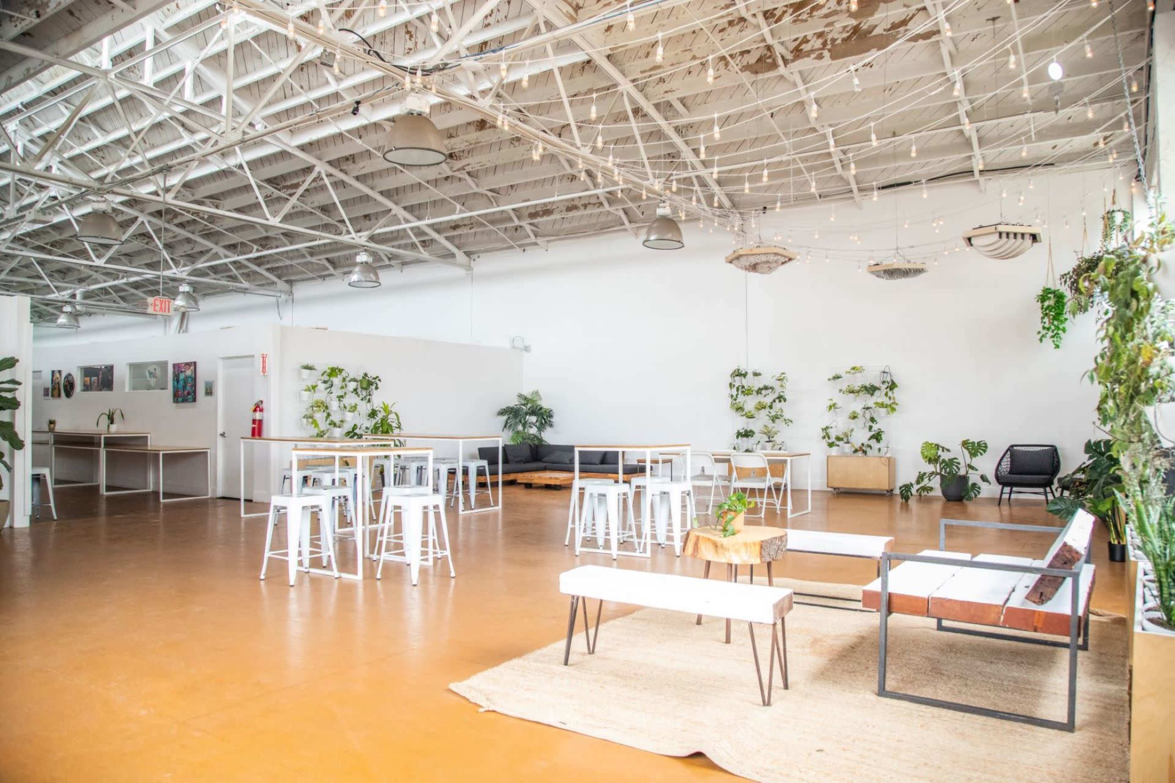 A spacious interior with several tables and chairs, a seating area, and various plants against white walls and a high ceiling.