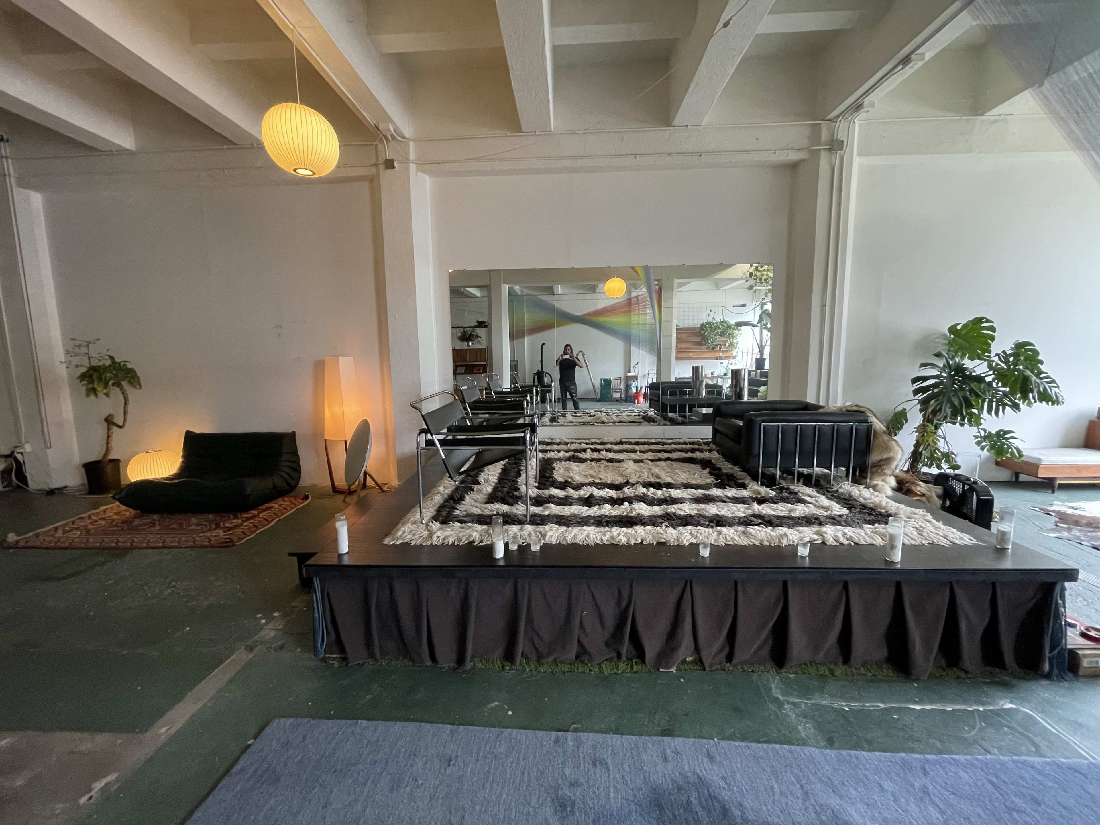A spacious indoor area with a raised platform covered in a patterned rug, surrounded by seating and plants.