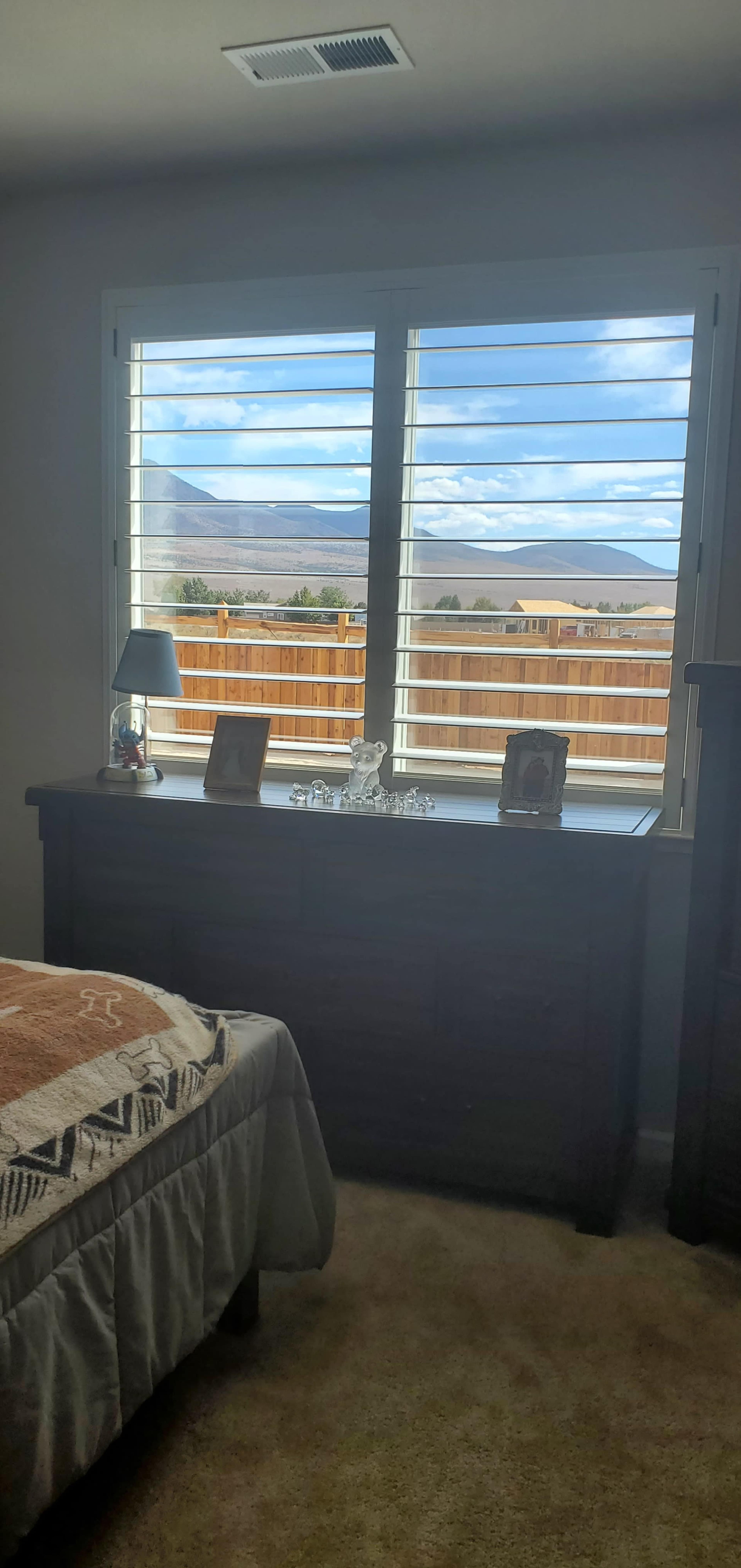 A bedroom with a neatly made bed, a wooden dresser beneath a window with plantation shutters, and a view of a fenced outdoor area and mountains.