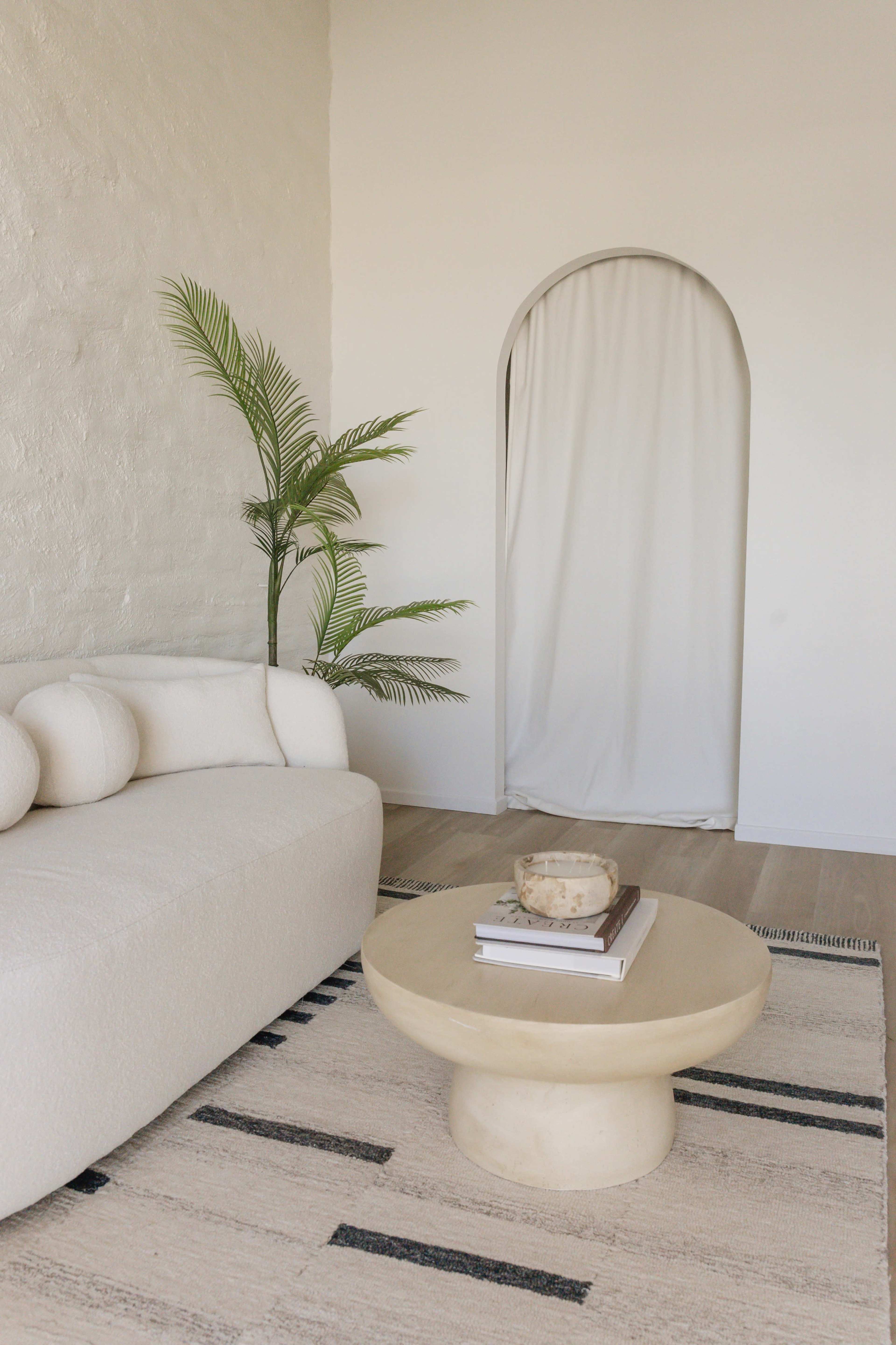 A light-filled living room features a textured white sofa, a round coffee table with stacked books, and a potted plant beside a softly draped archway.