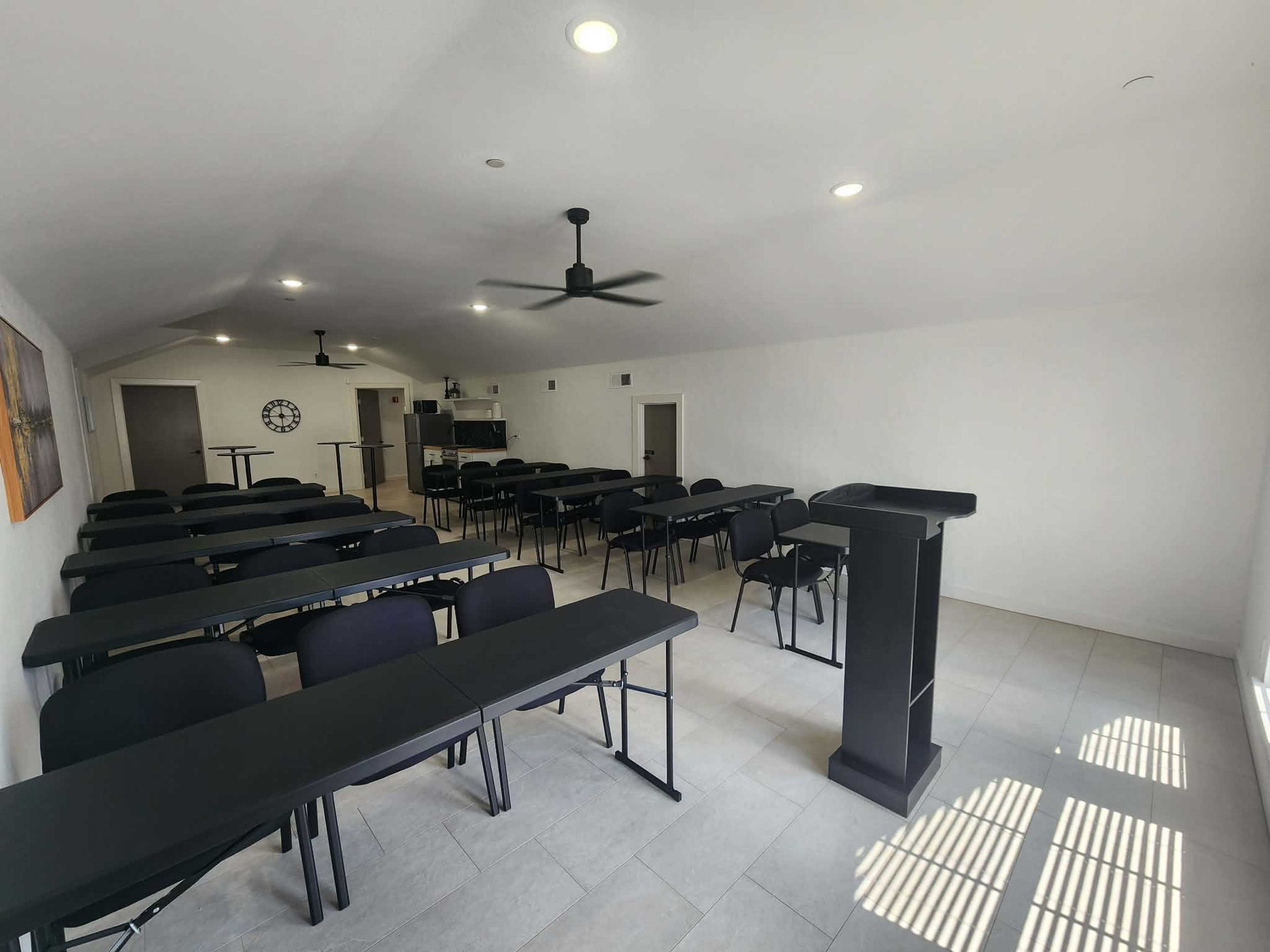 The image shows an empty classroom with rows of black tables and chairs, ceiling fans, and a podium at the front, illuminated by natural light.