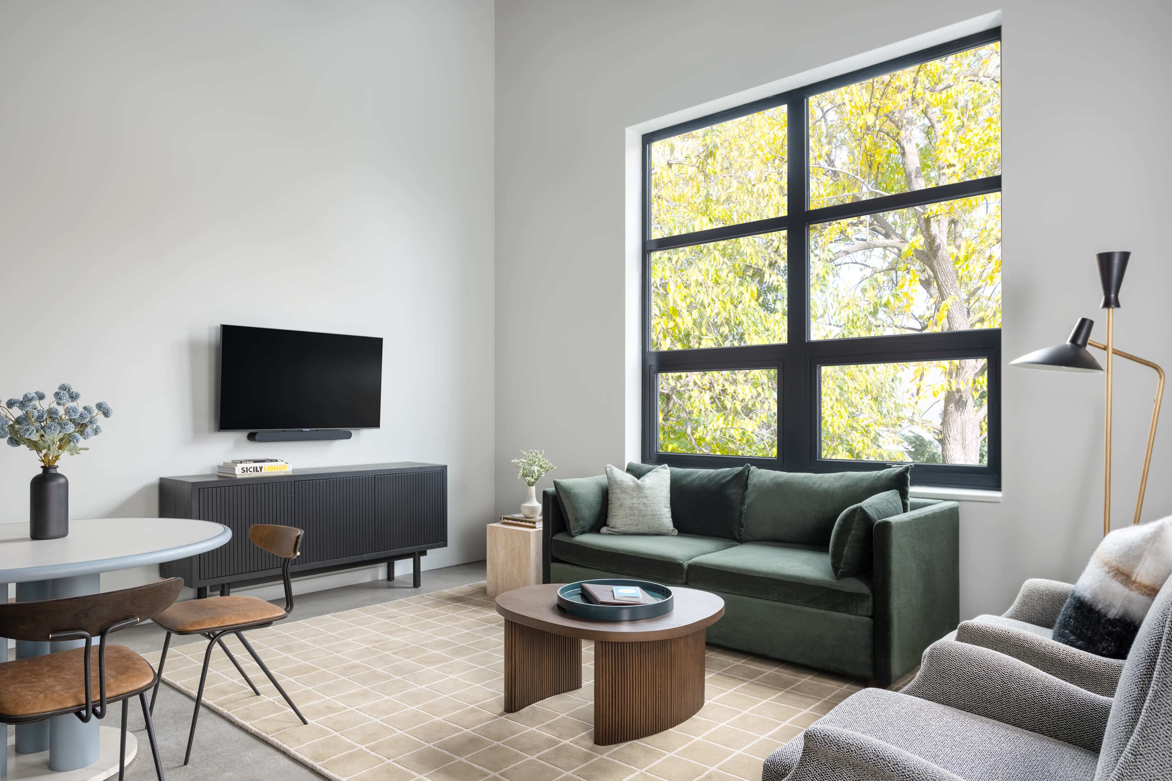 A modern living room features a green sofa, a round table with wooden chairs, and large windows with views of trees, all arranged around a coffee table on a carpet.