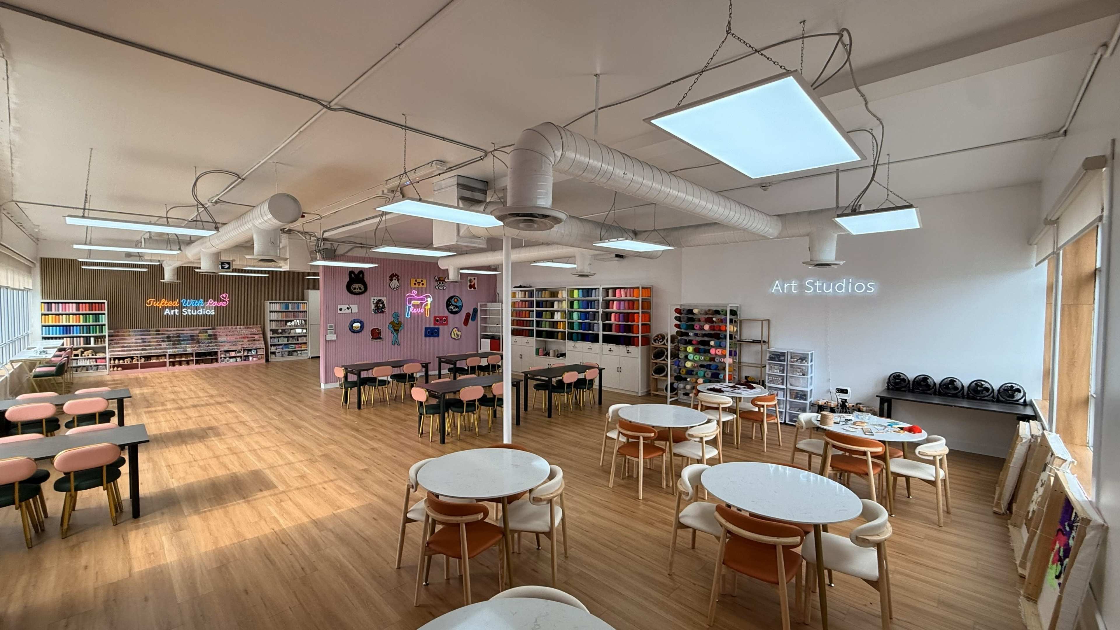 The image shows a brightly lit art studio with tables, chairs, and colorful shelves filled with art supplies along the walls.