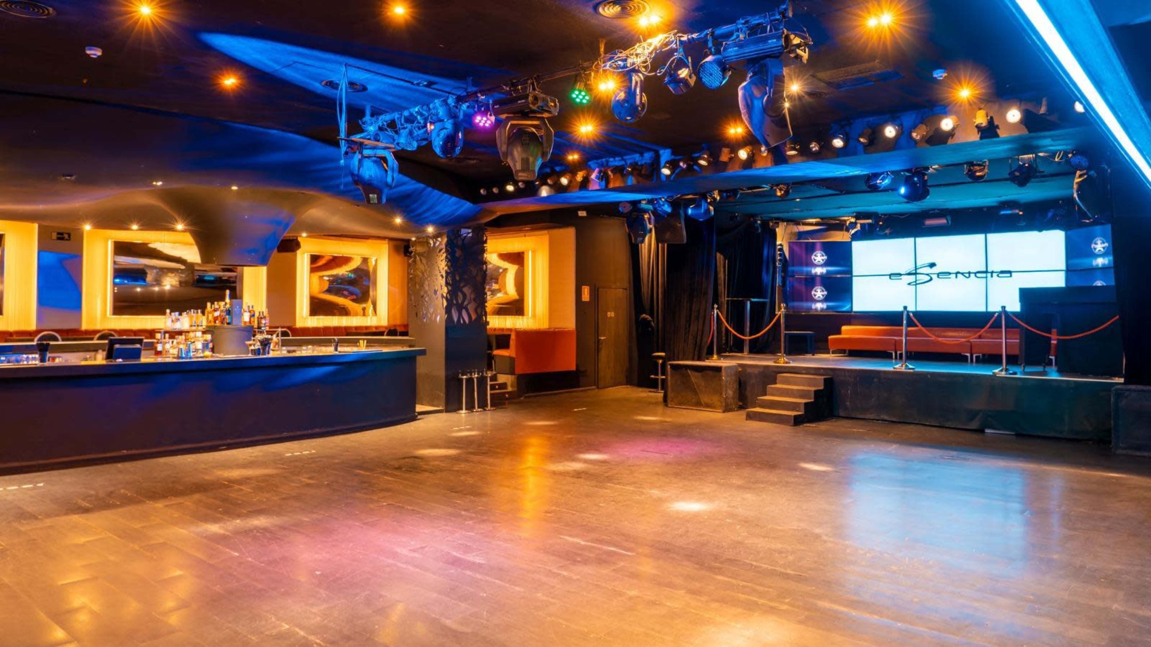 The image shows an empty nightclub interior featuring a bar area, a stage with lighting, and a dance floor.