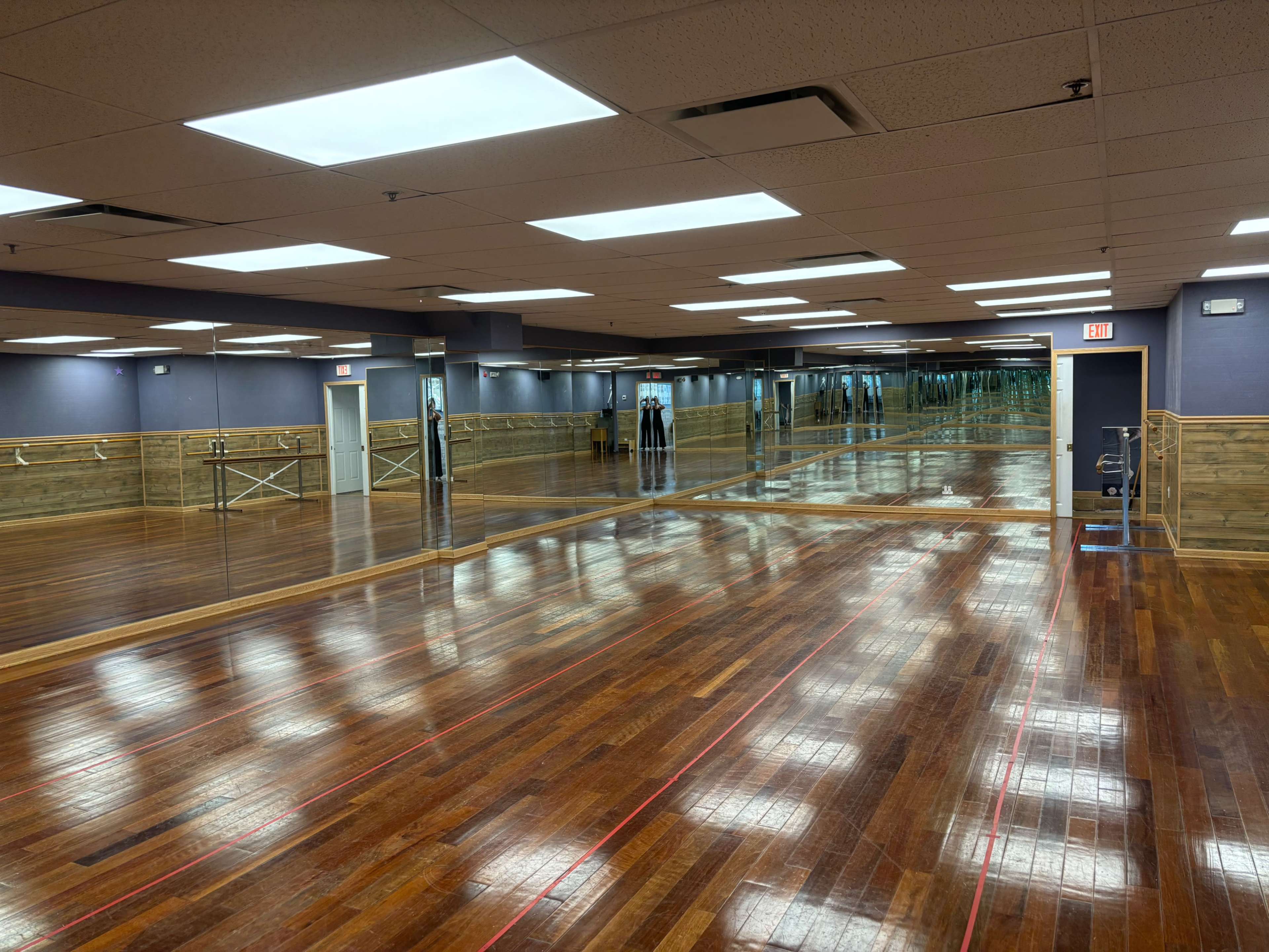 A spacious dance studio with wooden floors, mirrored walls, and bright overhead lighting.