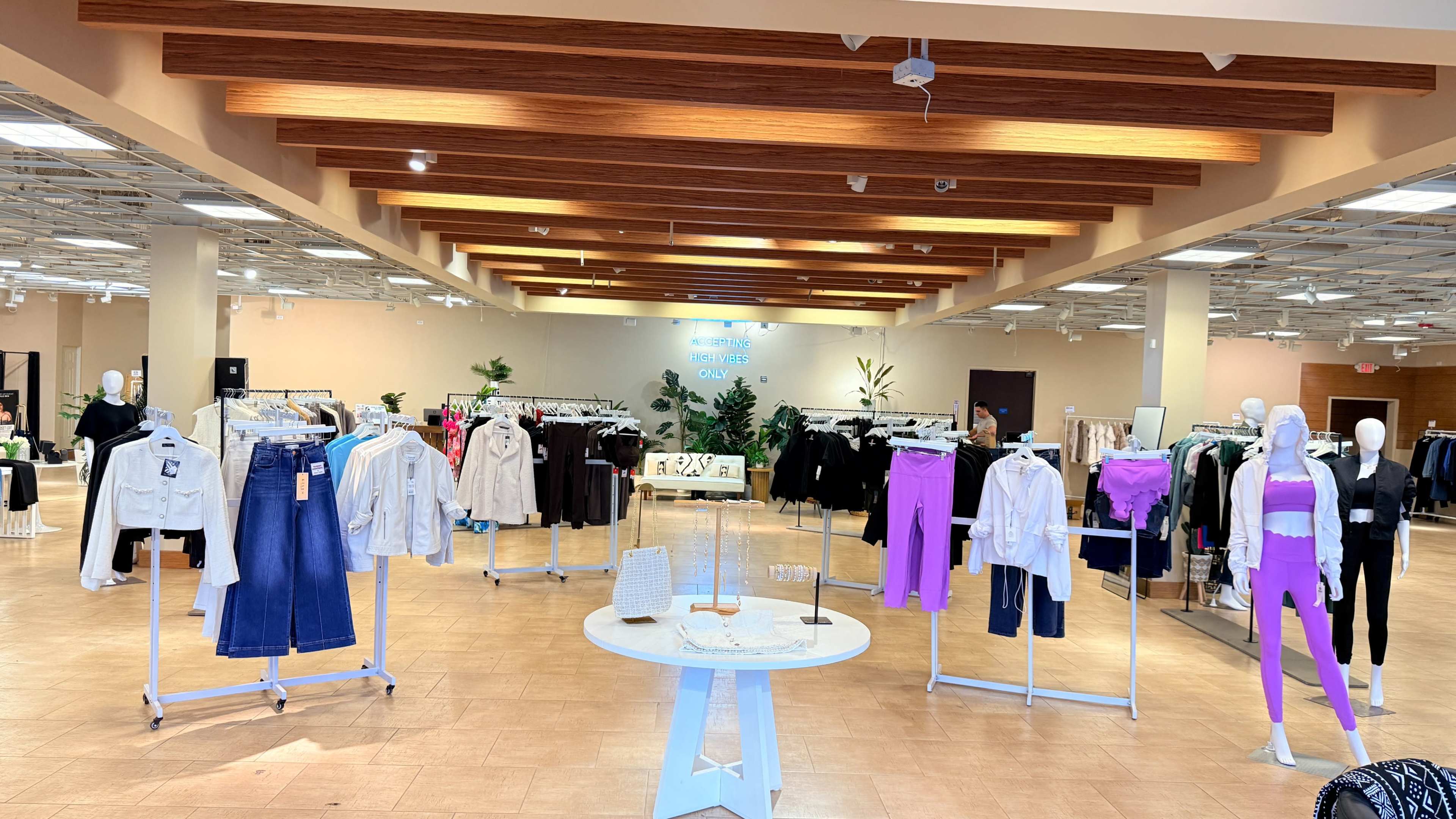 The image shows a spacious retail clothing store with rows of apparel on racks, including various tops and bottoms, under wooden beams and bright lighting.