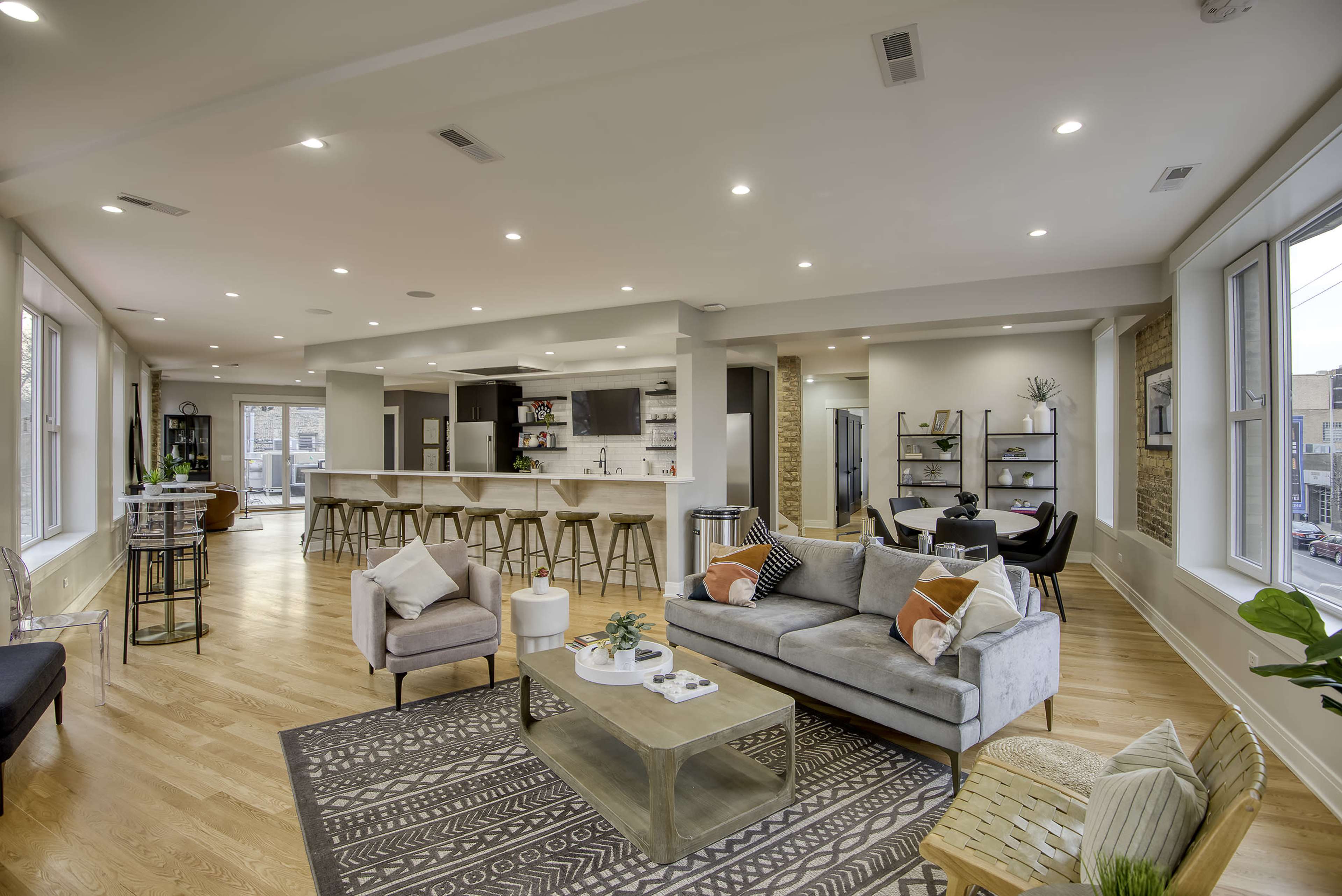 A spacious modern living area with a gray sofa, a glass coffee table, a dining area, and large windows allowing natural light to fill the space.