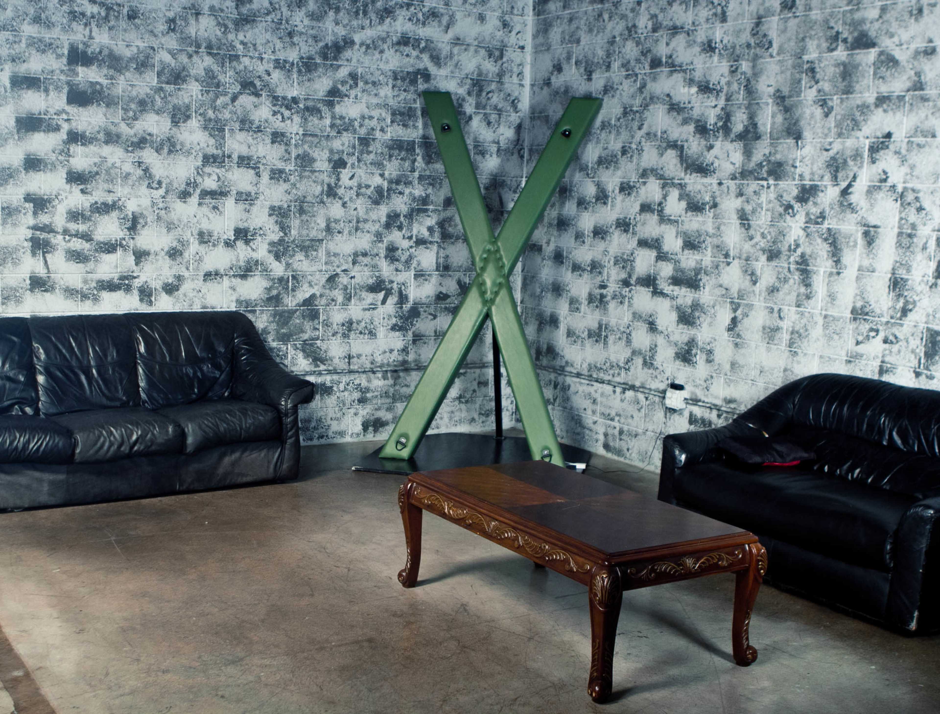 A minimalist room with two black leather sofas and a wooden coffee table, featuring a large green X-shaped support beam on the wall.