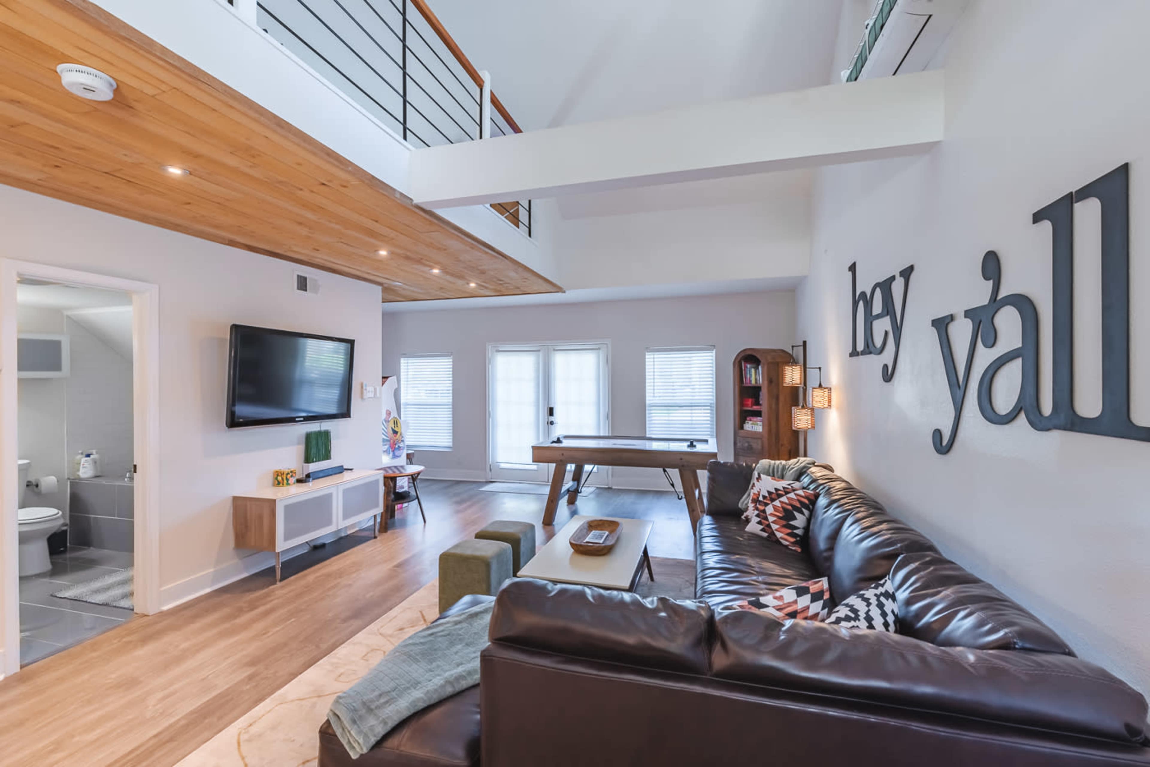 The image shows a spacious living area with a brown leather sofa, a large wooden table, a television on the wall, and large windows letting in natural light.