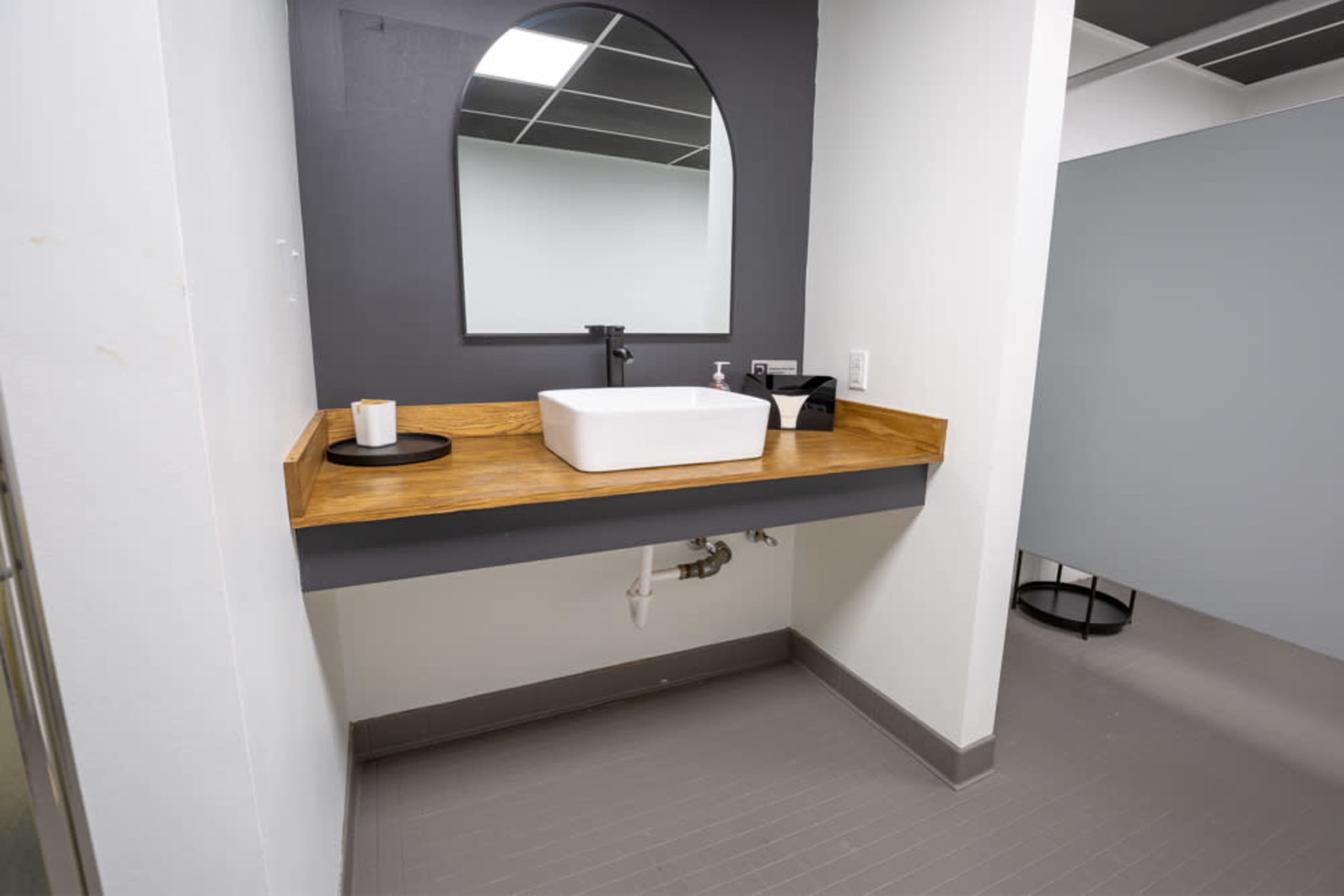 The image shows a modern bathroom with a square white sink on a wooden countertop, a wall-mounted faucet, and a mirror in an arched frame.