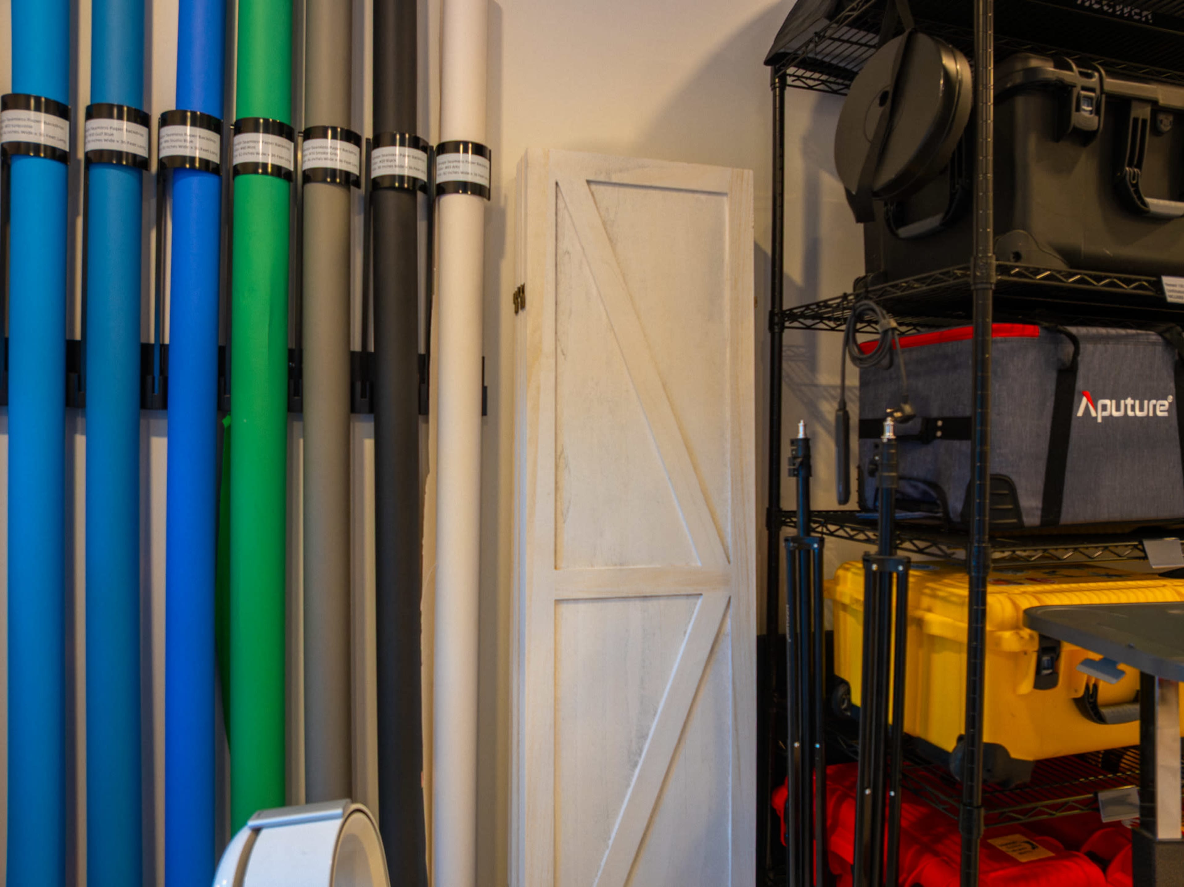 The image shows a room with various colored backdrop rolls leaning against a wall and a storage unit holding photography equipment and supplies.