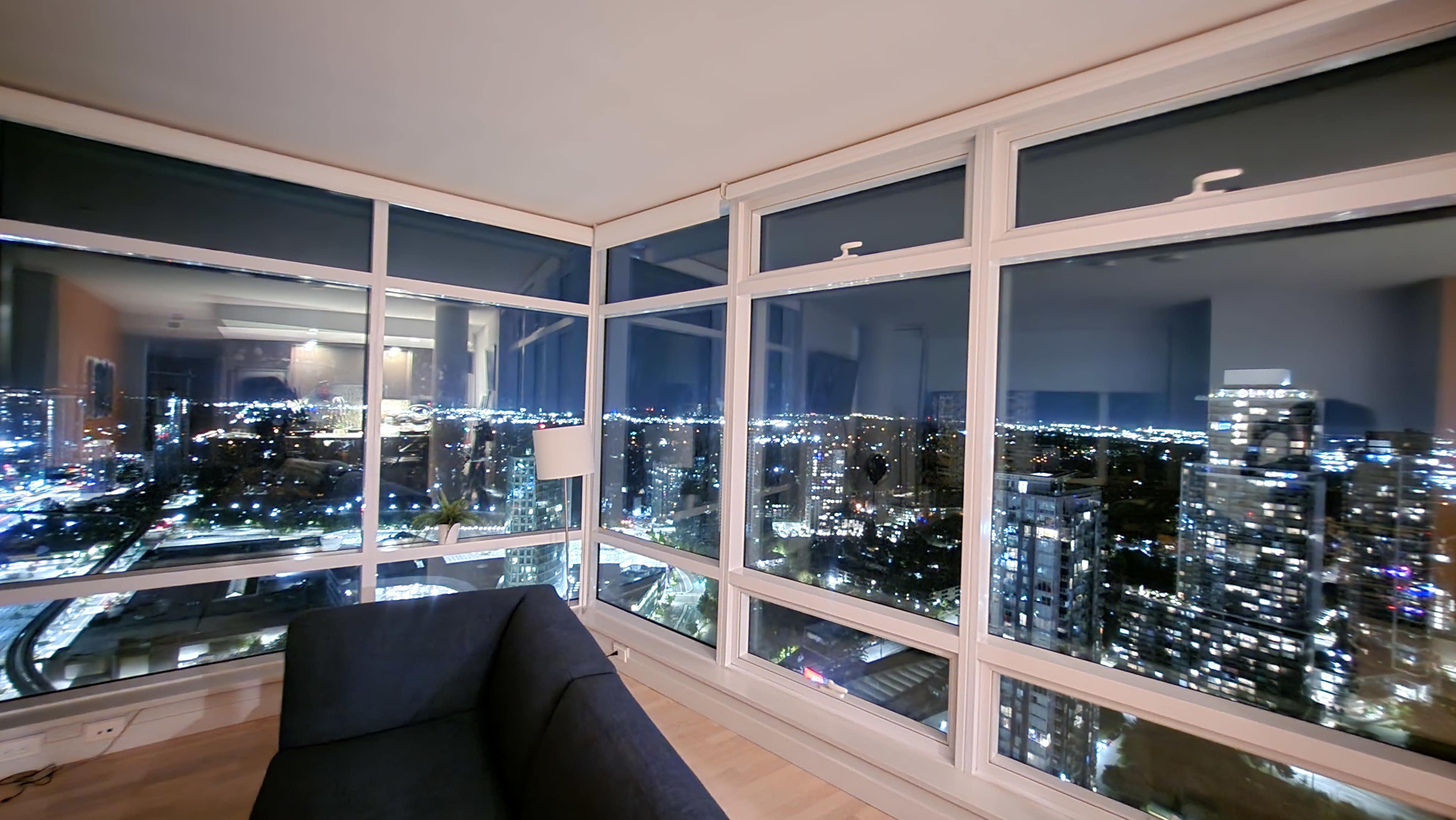 A modern apartment features large glass windows offering a panoramic view of a city skyline at night, illuminated by city lights.