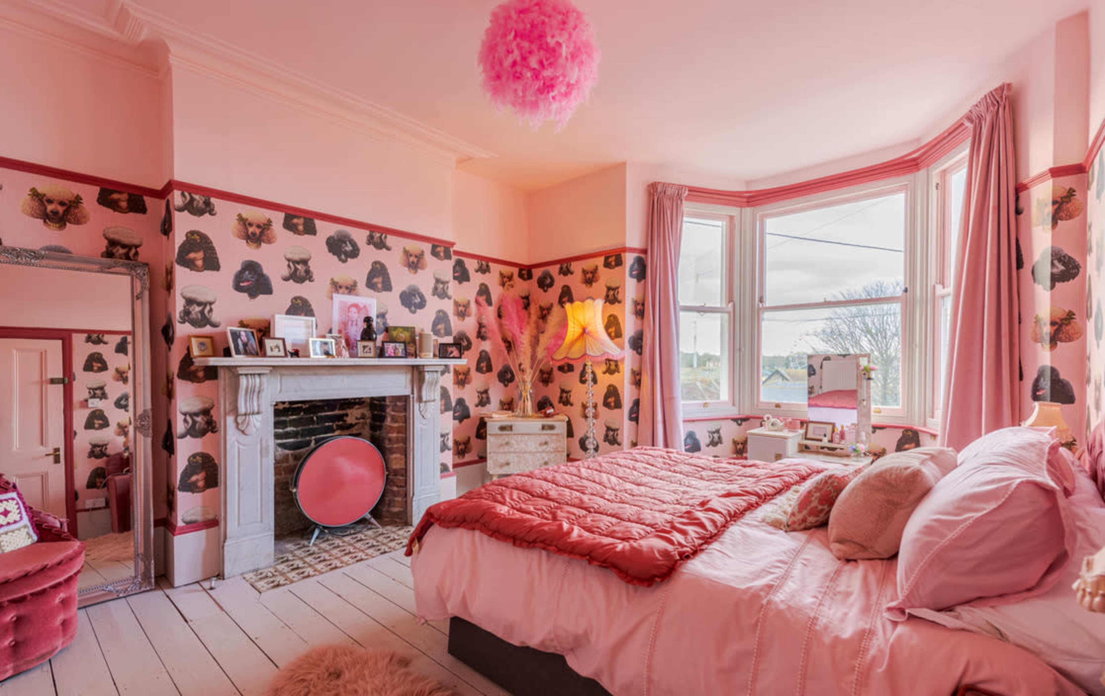 A pink-themed bedroom featuring patterned wallpaper, a plush bed with a red cover, and a fireplace adorned with decorative items.