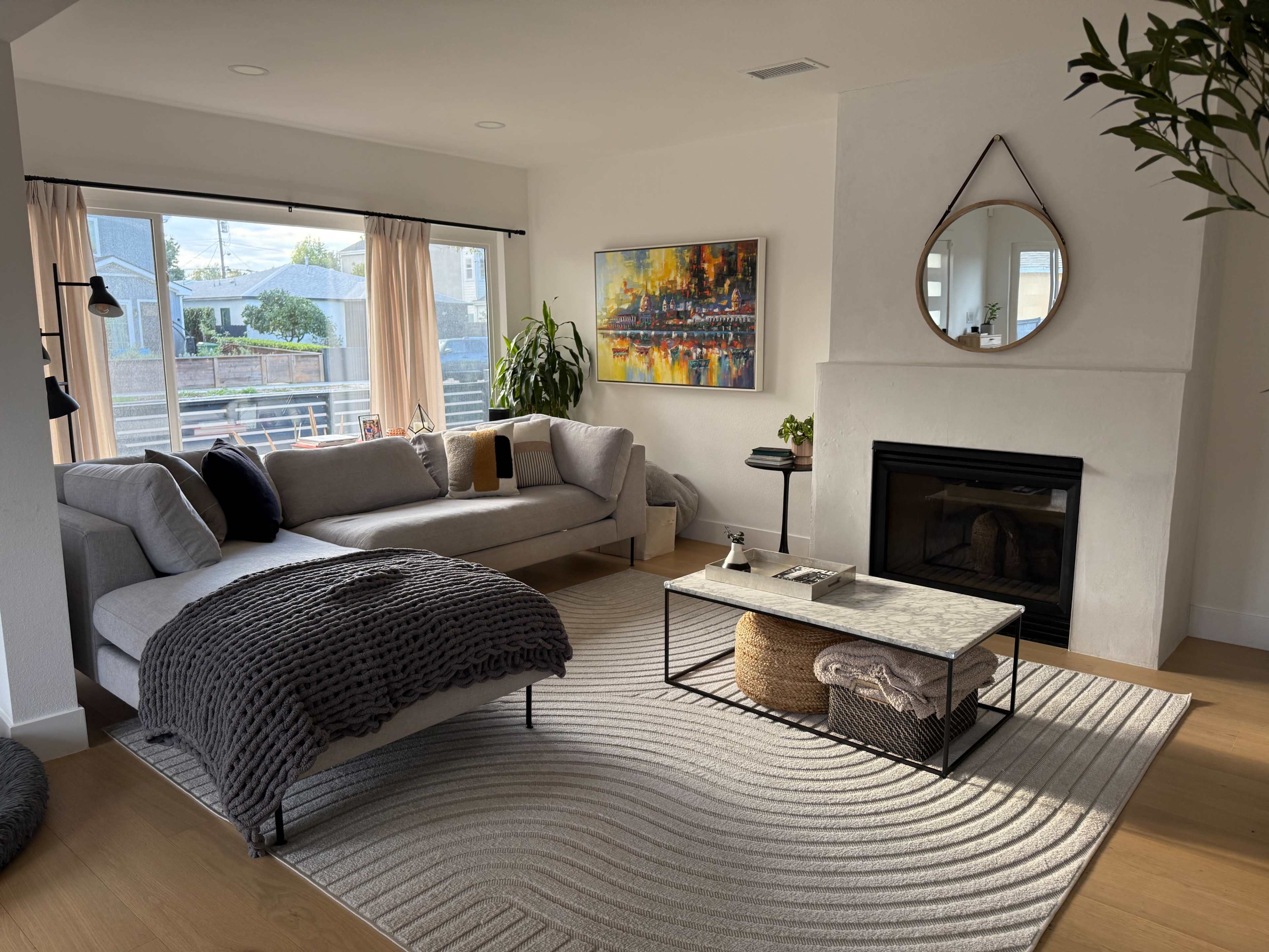 The image shows a modern living room featuring a gray sectional sofa, a coffee table with a textured surface, and large windows with natural light illuminating the space.