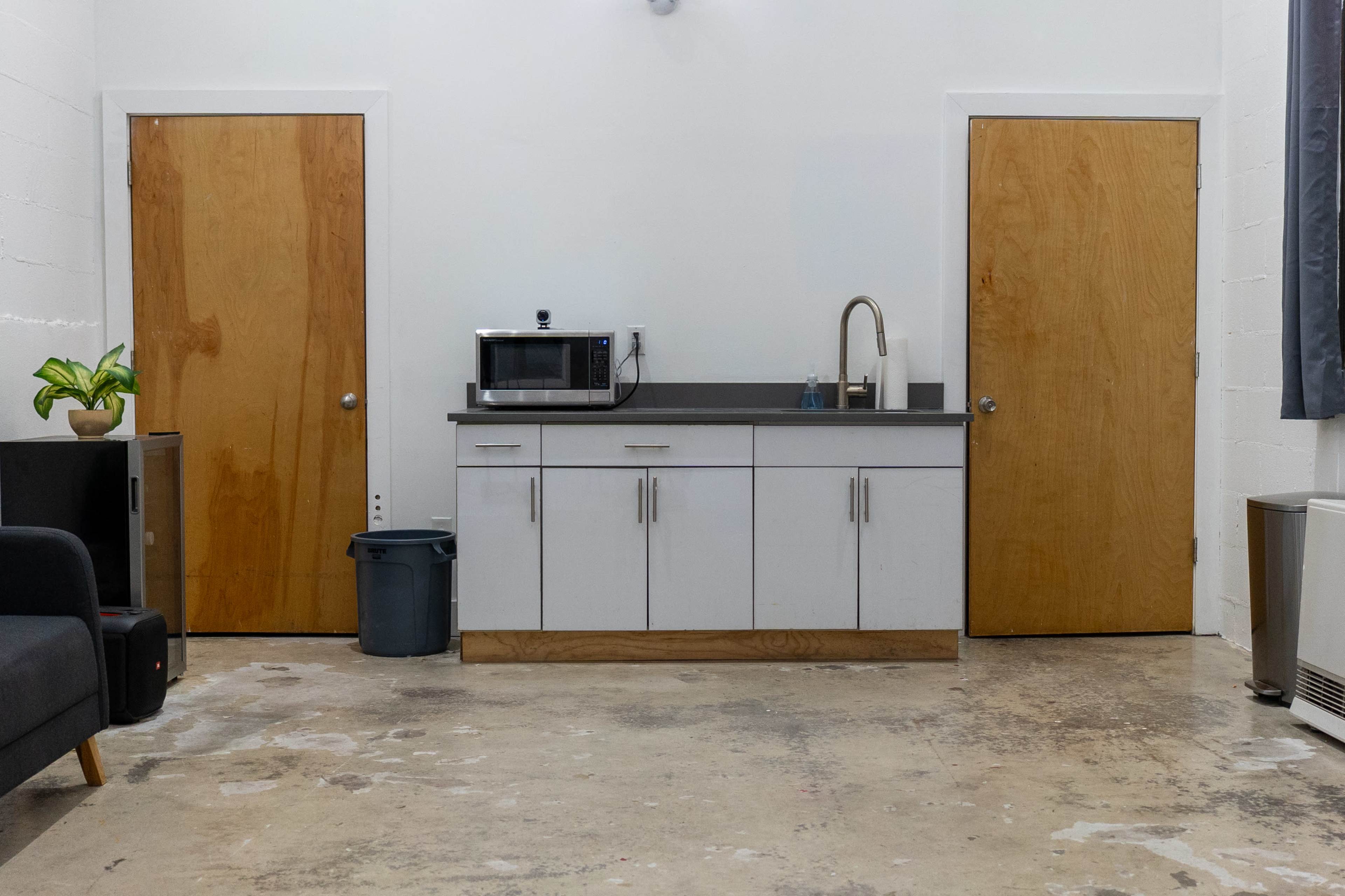 A minimalist kitchen area with a microwave, sink, and two wooden doors on either side, set against a concrete floor.