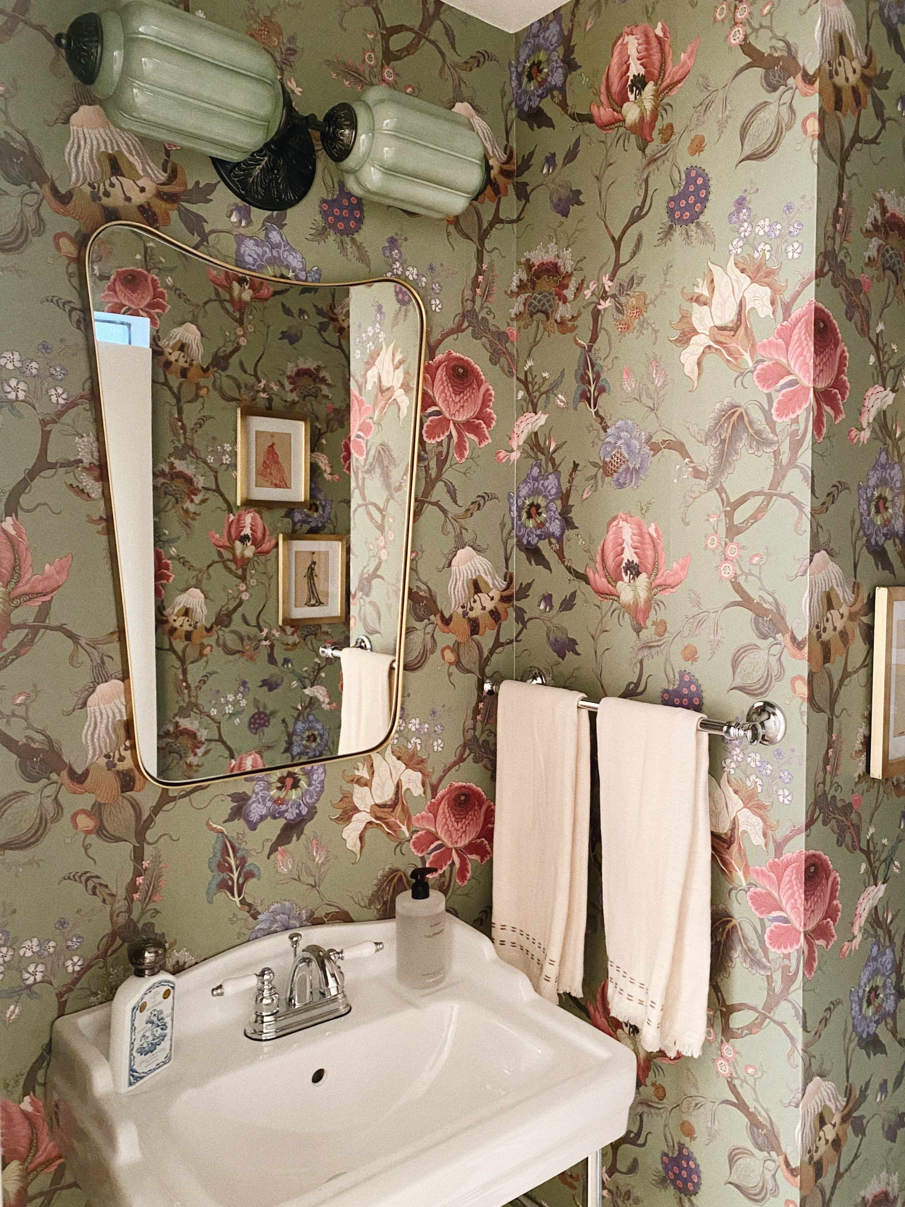 A small bathroom features floral wallpaper, a curved mirror, and a white sink with two towels hanging beside it.