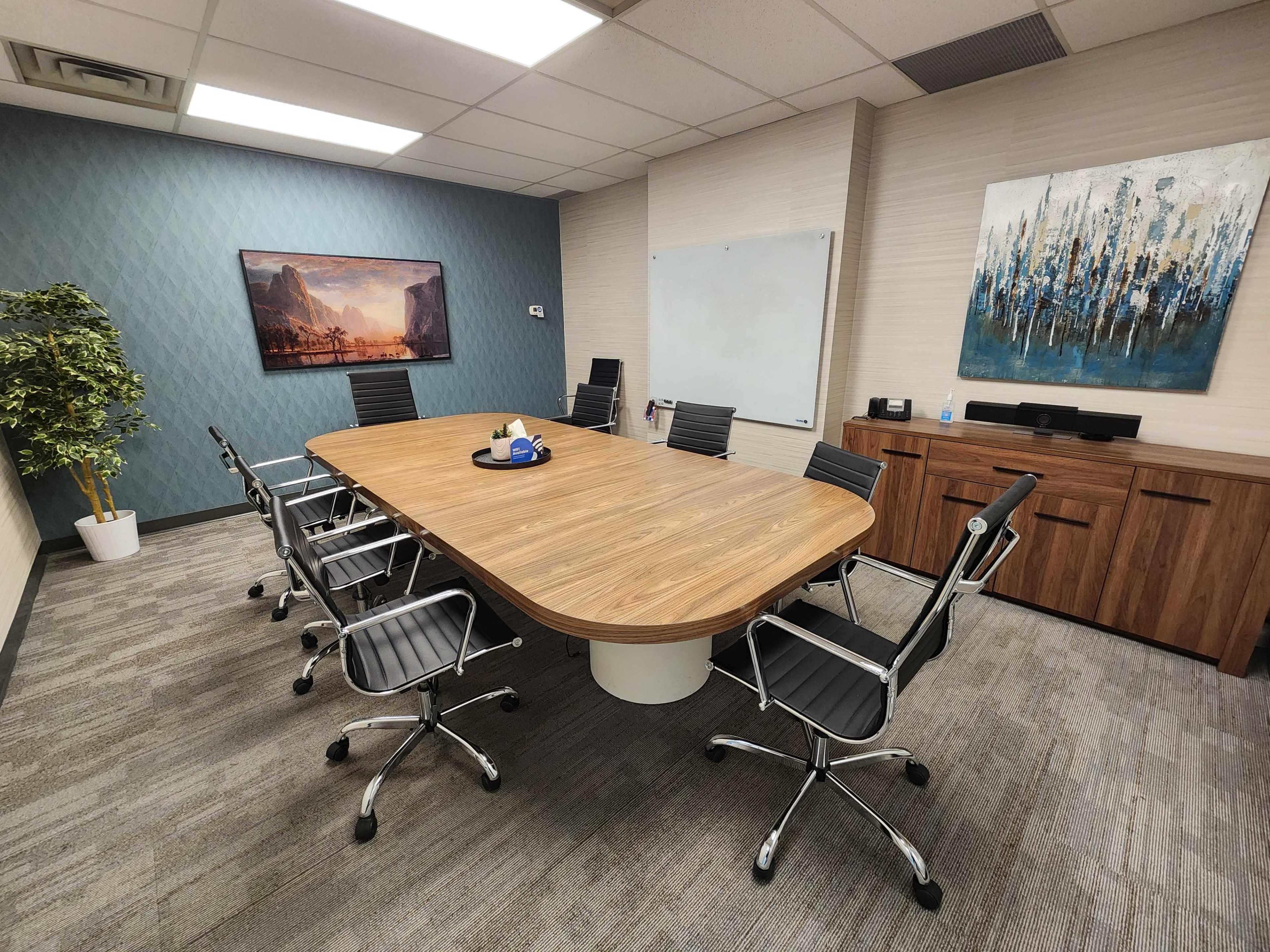 A modern conference room features a large oval wooden table surrounded by black rolling chairs, with artwork and a whiteboard on the walls.
