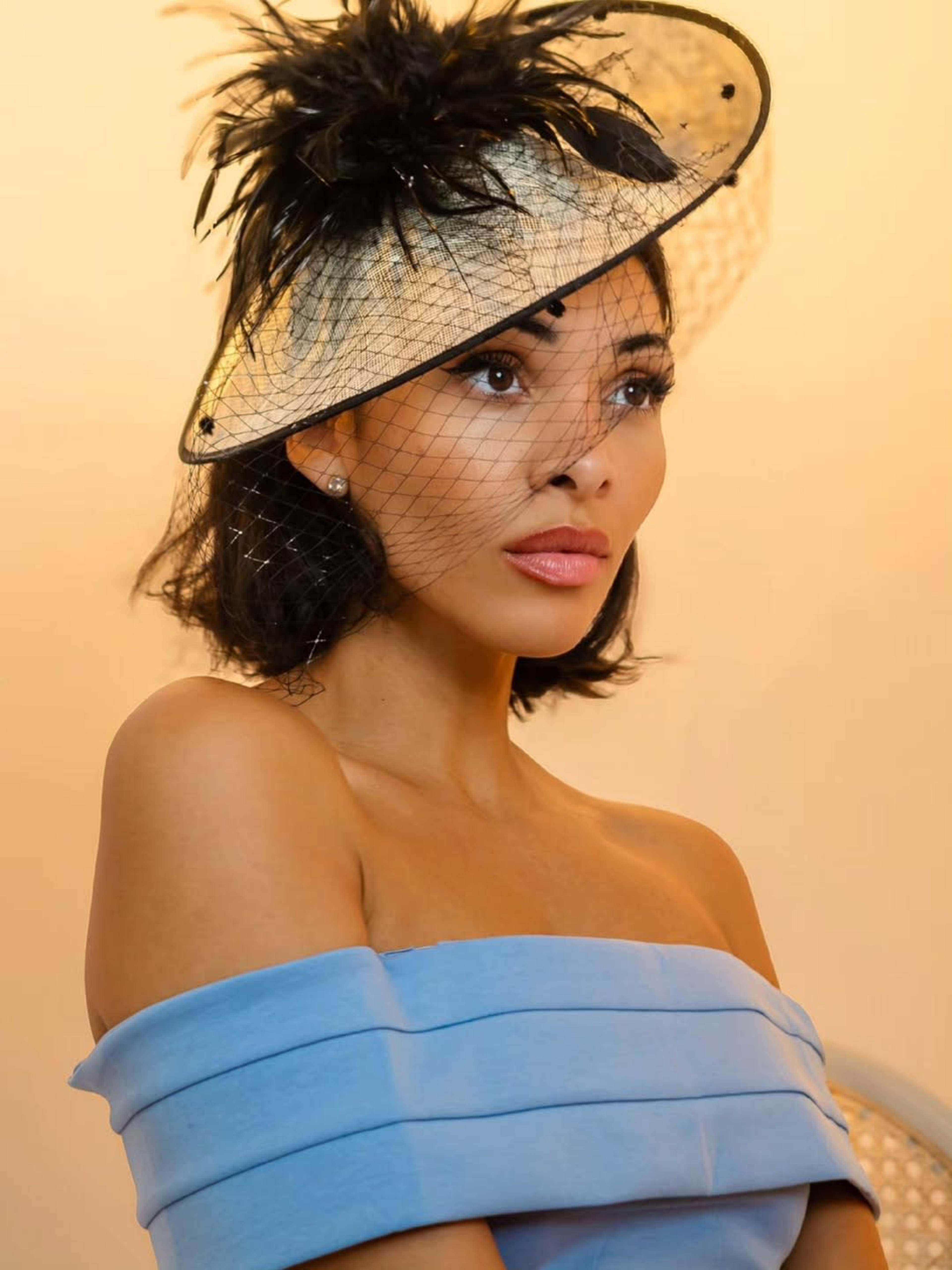 The image features a woman wearing a structured blue off-shoulder dress and a large, decorative black hat with a netting detail.
