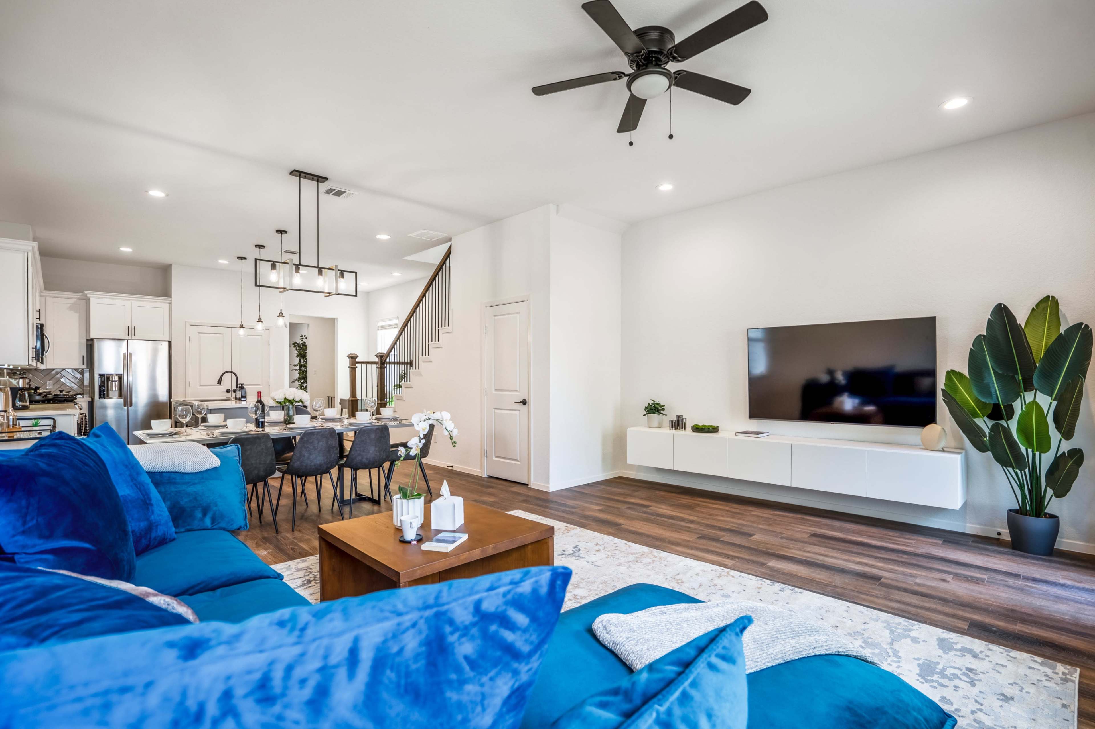 A modern living room features a blue sectional sofa, a wooden coffee table, and a dining area with a large table and chairs, connected to a kitchen with white cabinetry.