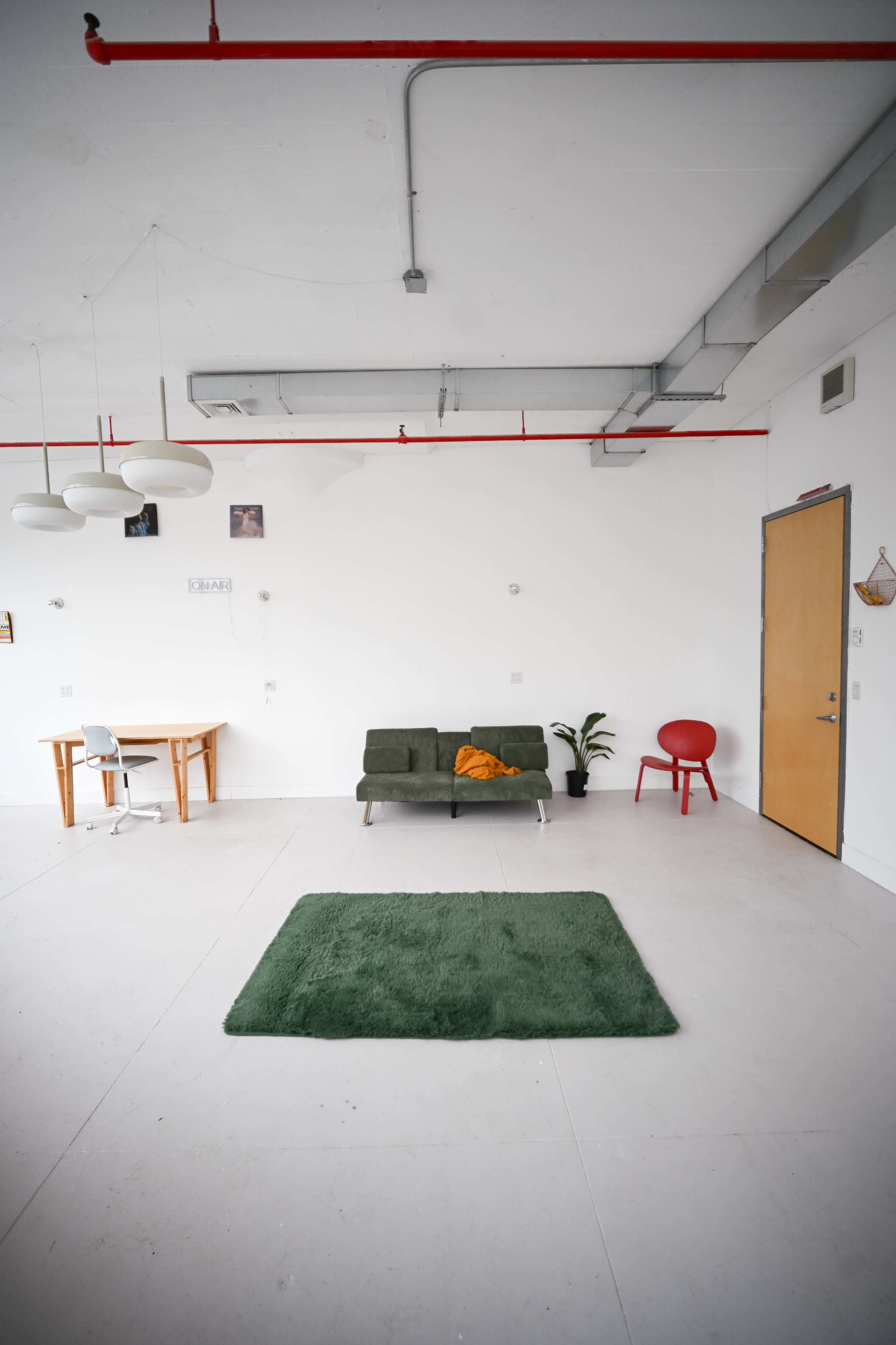 The image shows a minimalistic, brightly lit room featuring a green rug, a green sofa with an orange throw, a wooden dining table, and a red chair against white walls.