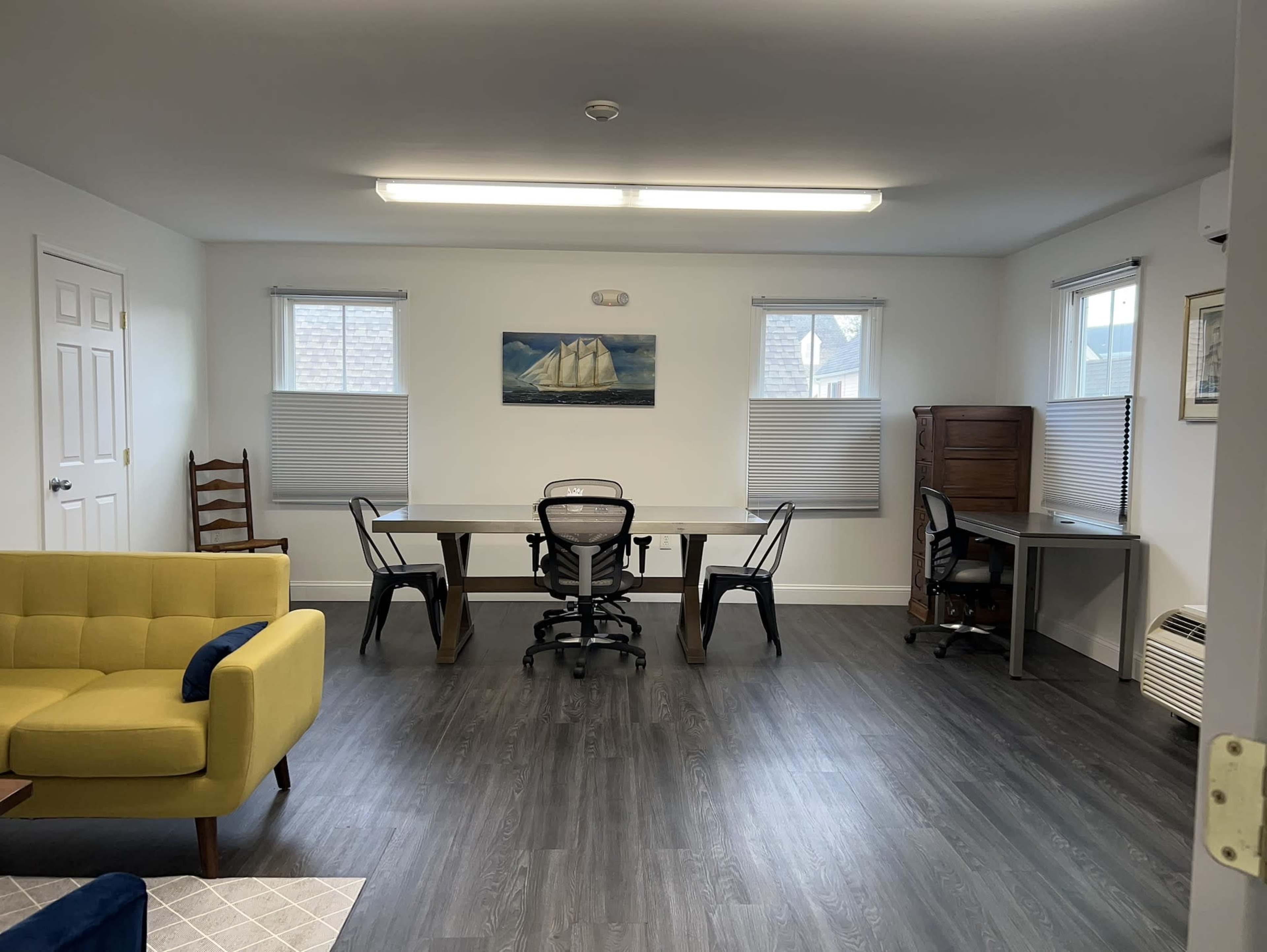The image shows a bright room with a yellow couch, two desks, several chairs, and windows with blinds.