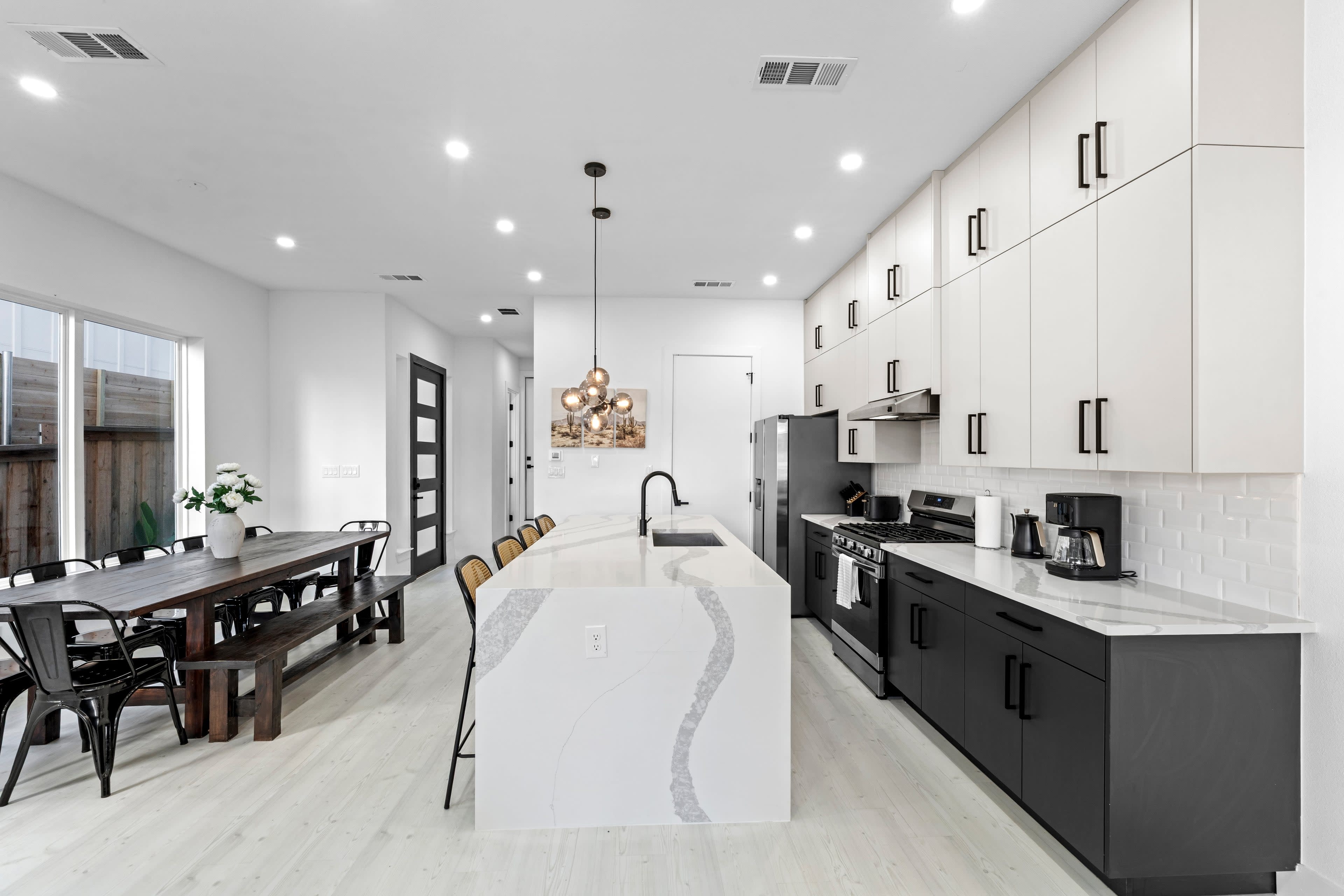 The image shows a modern kitchen featuring a large island with a white countertop, black lower cabinets, and stainless steel appliances, alongside a dining area with a wooden table and metal chairs.