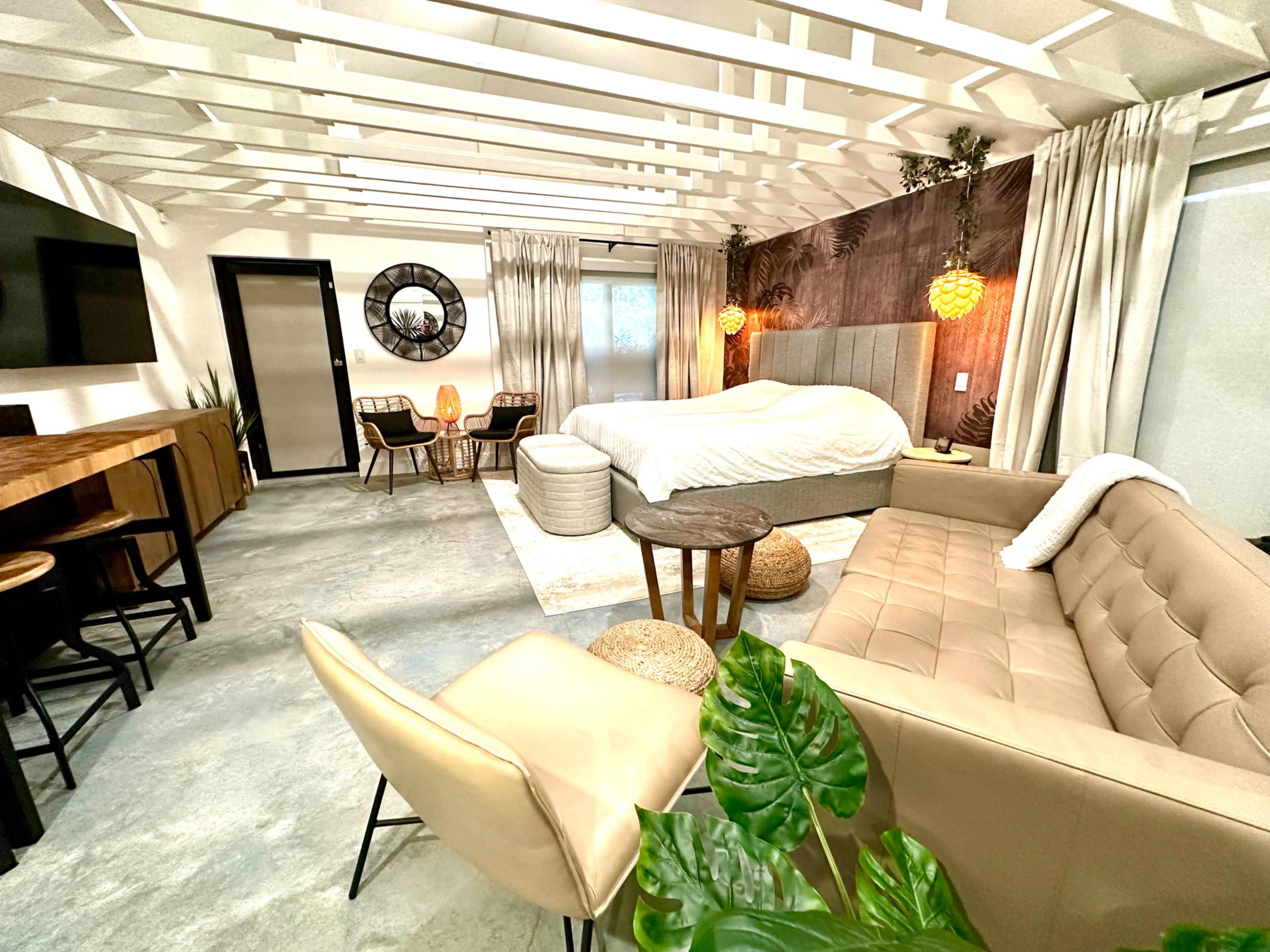 A modern bedroom featuring a large bed, a couch, a dining area with bar stools, and decorative plants against a backdrop of light-colored walls and wood accents.