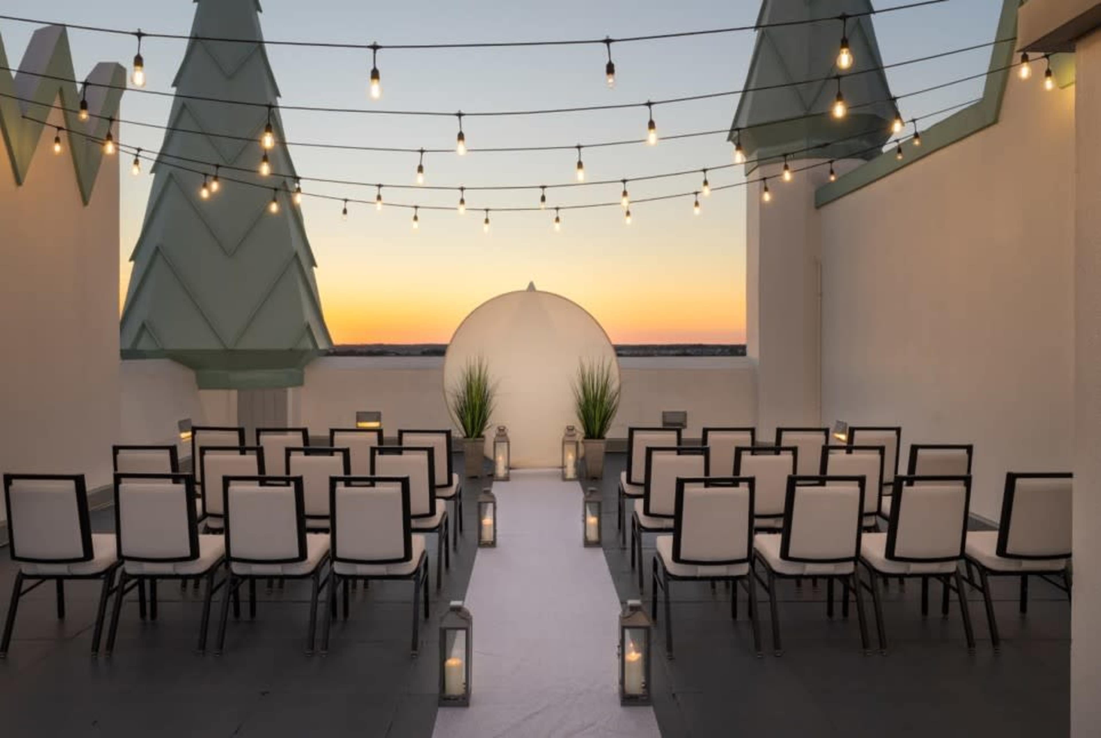 A rooftop venue is set up for a ceremony, featuring rows of empty chairs, lanterns lining the aisle, and string lights overhead against a sunset backdrop.