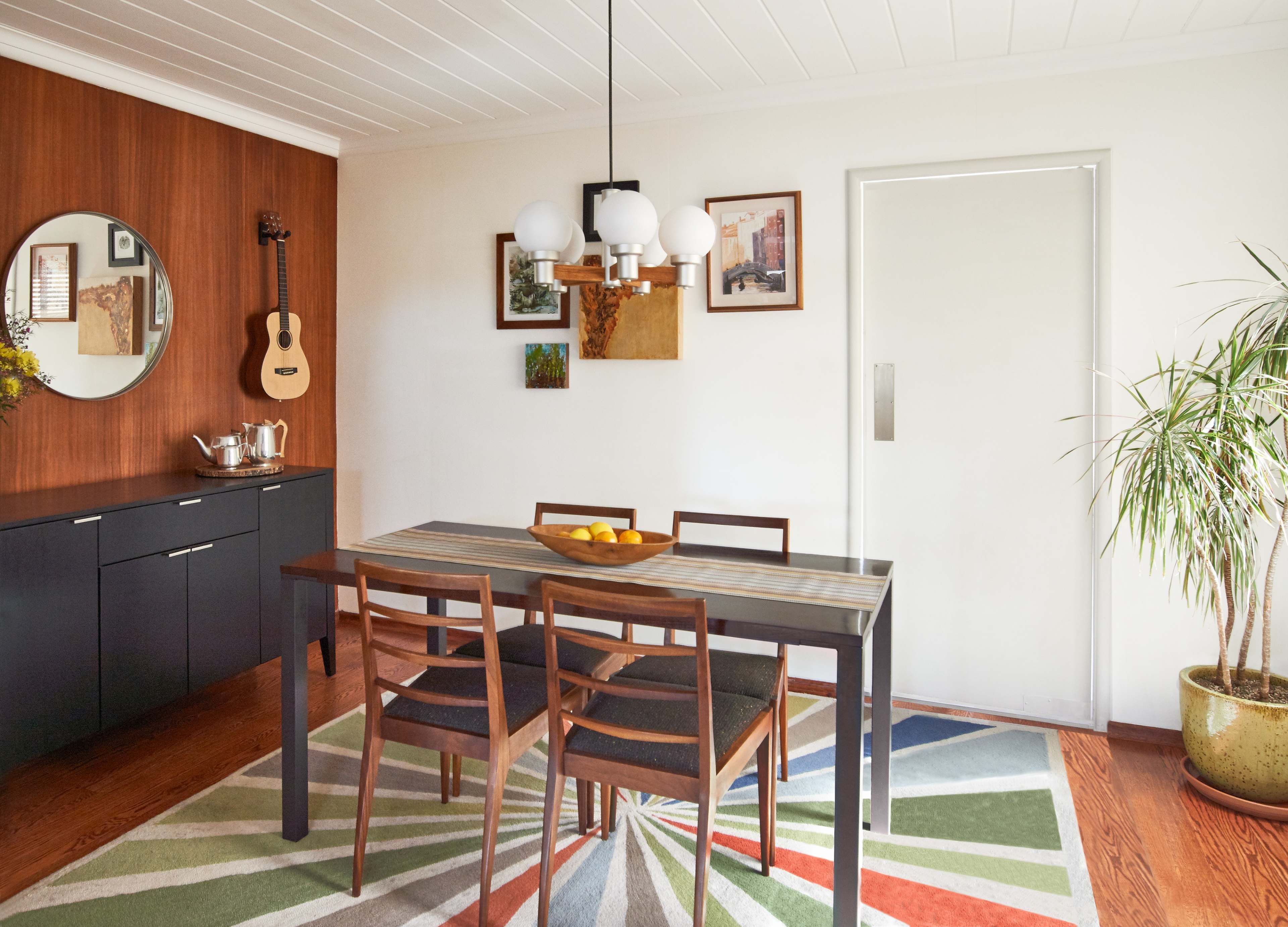 A modern dining area features a dark wooden table with four chairs, a geometric rug, and art on the walls, alongside a door and a potted plant.