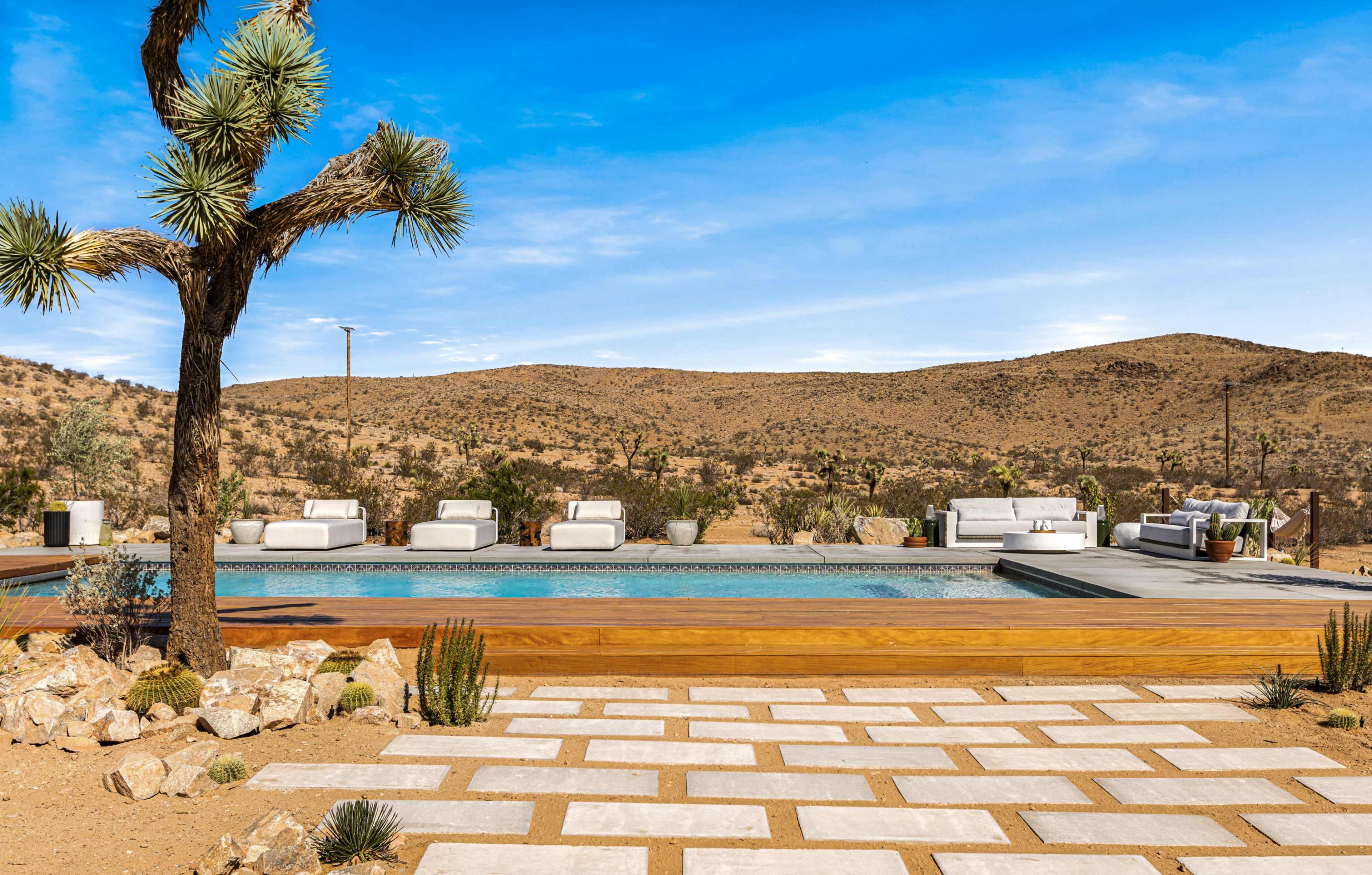 A desert landscape with a pool surrounded by white lounge chairs, set against rolling hills and a clear blue sky.