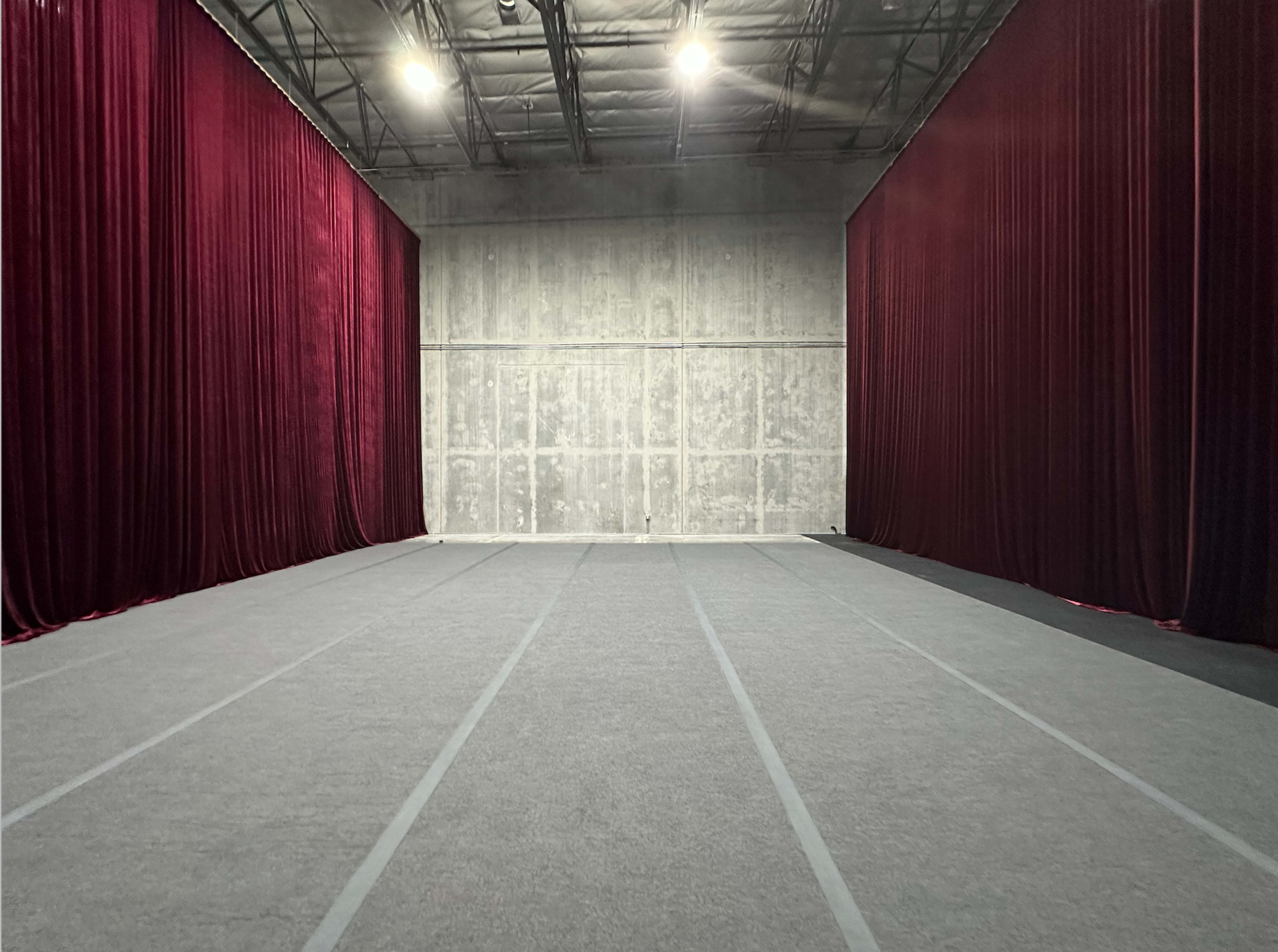 The image shows an empty space divided by deep red curtains, with a gray carpet and concrete walls.