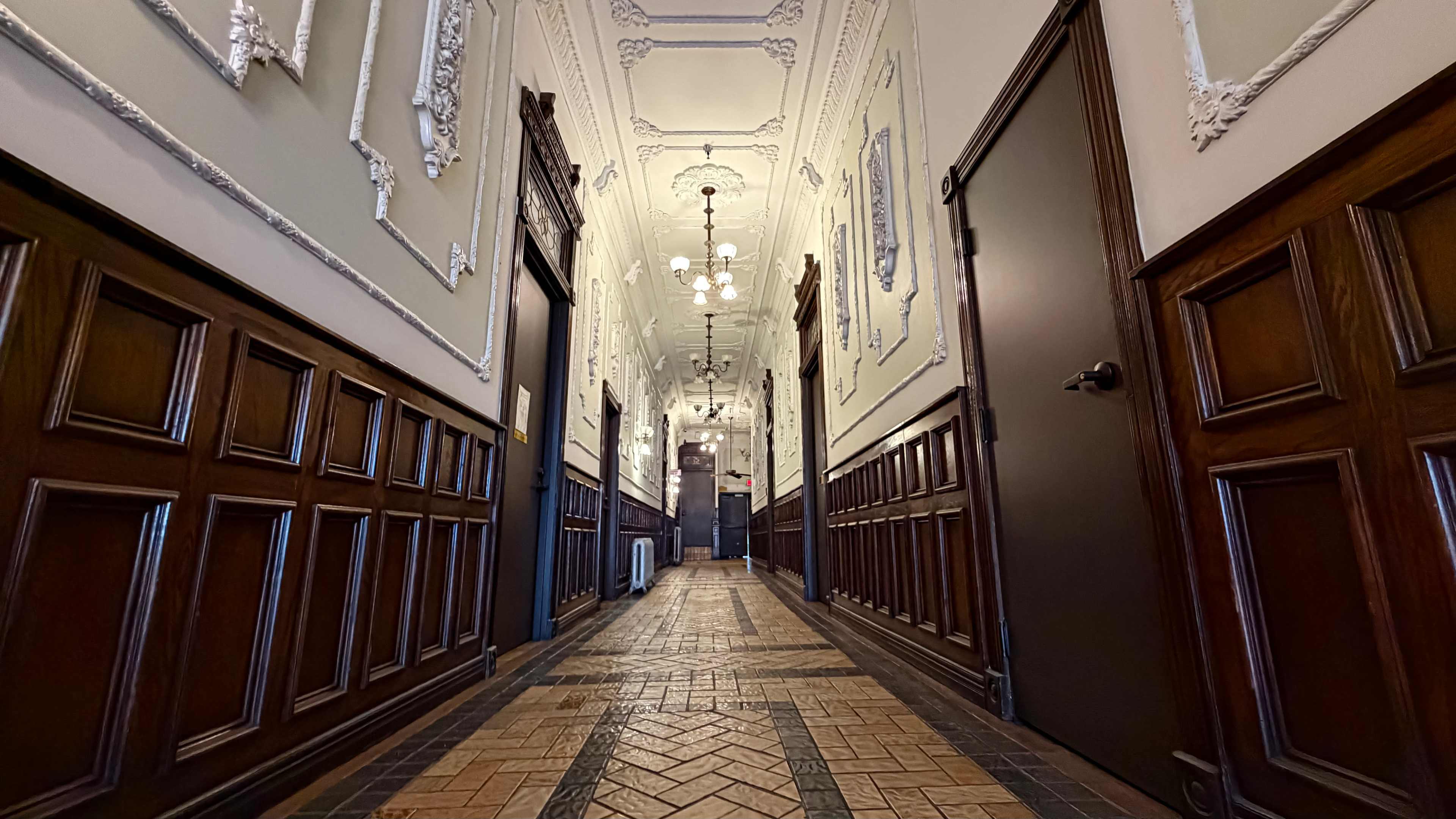 The image shows a long, ornate hallway with detailed wood paneling, decorative walls, and multiple doors leading to rooms on either side.