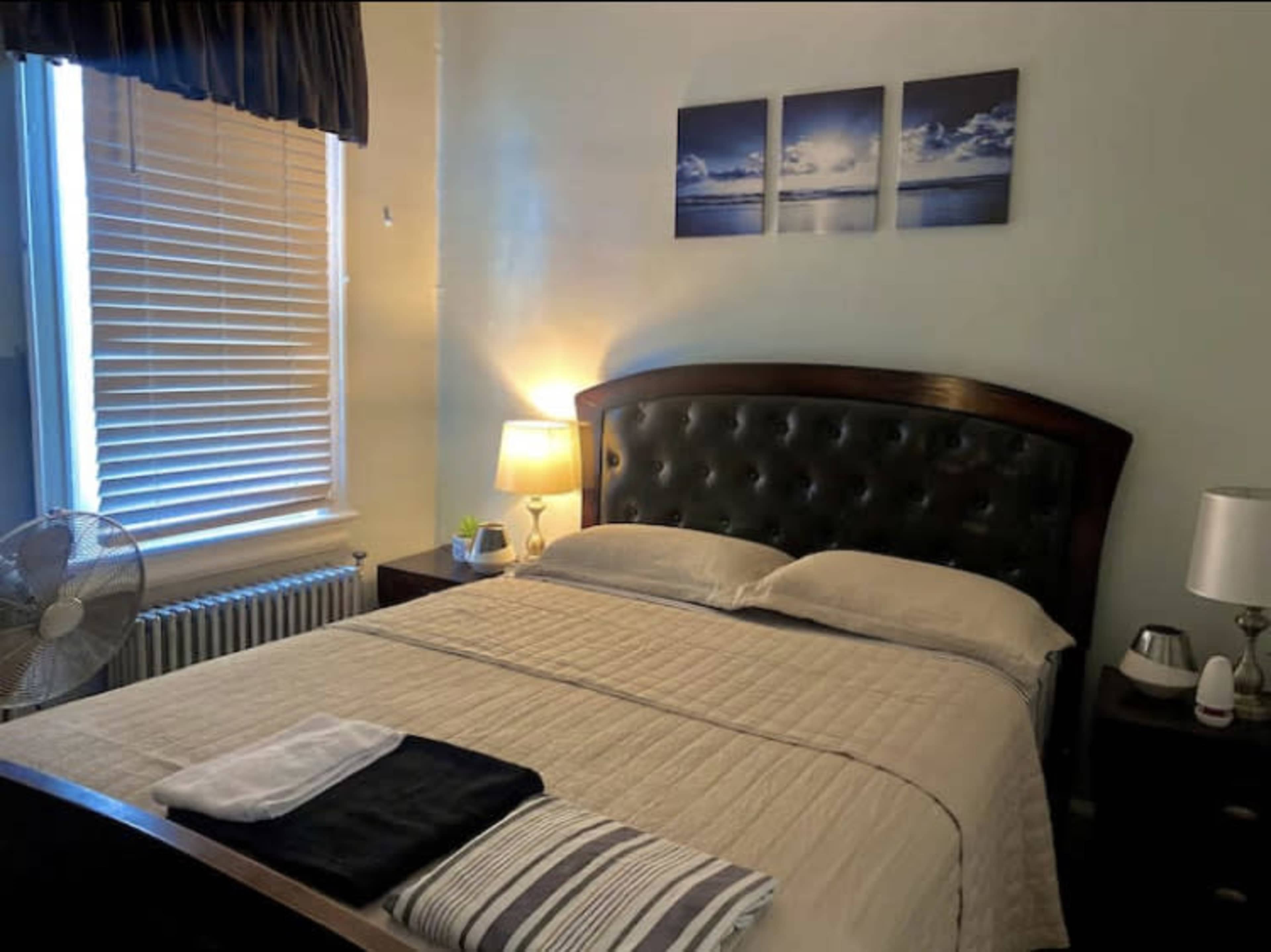 A neatly made bed with a beige quilt and decorative towels, framed by a bedside table with a lamp and a window with blinds.