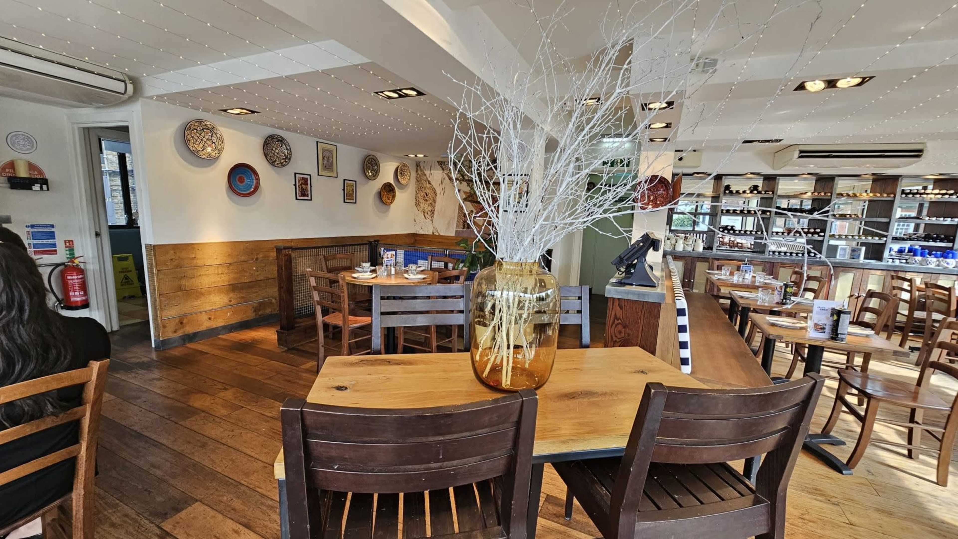 The image shows the interior of a restaurant with wooden tables, chairs, and decorative wall art, featuring a large vase with decorative branches at the center of a table.
