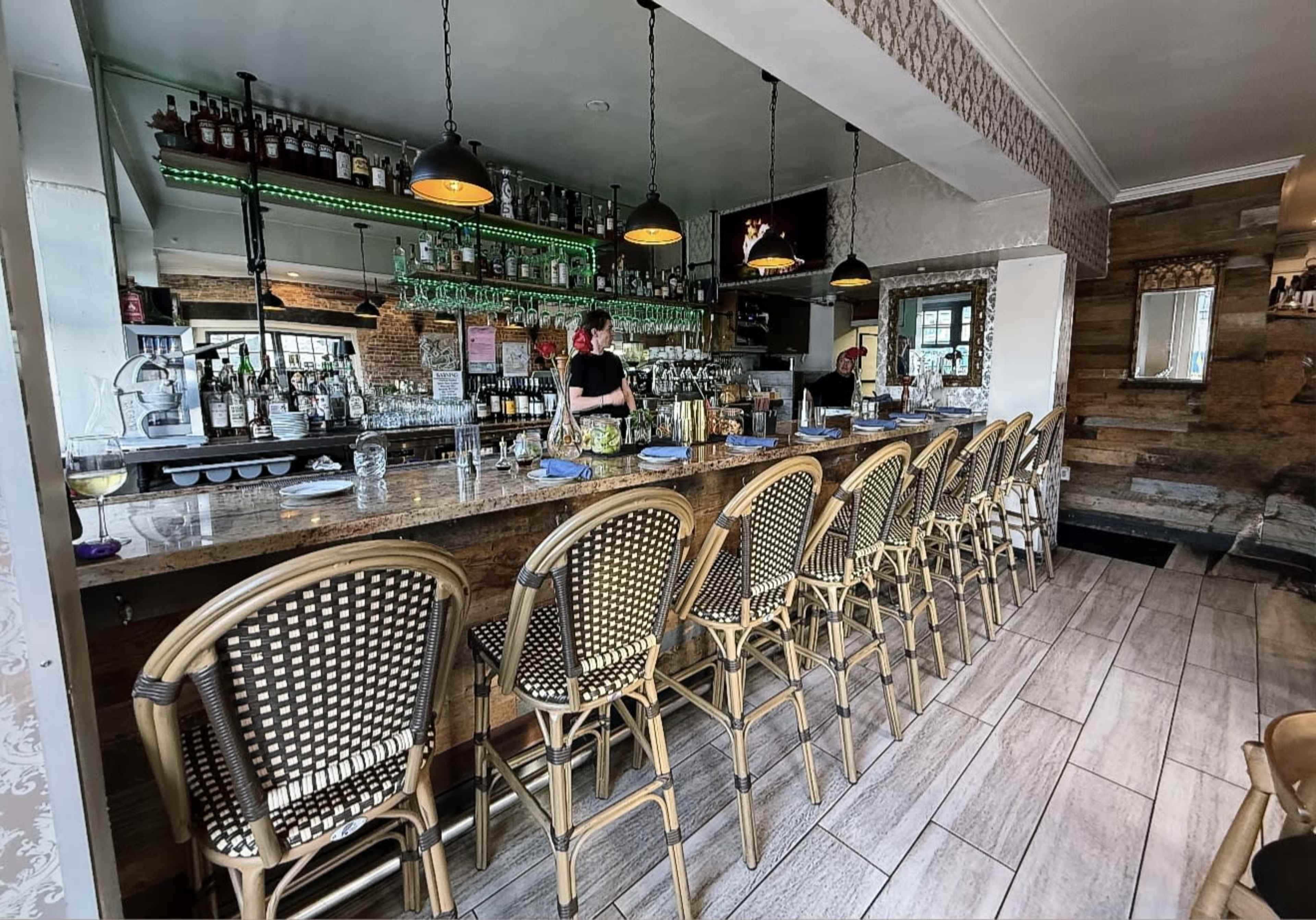 The image shows a bar with a long wooden counter and seated stools, where a bartender is preparing drinks behind a well-stocked shelf of liquor bottles.
